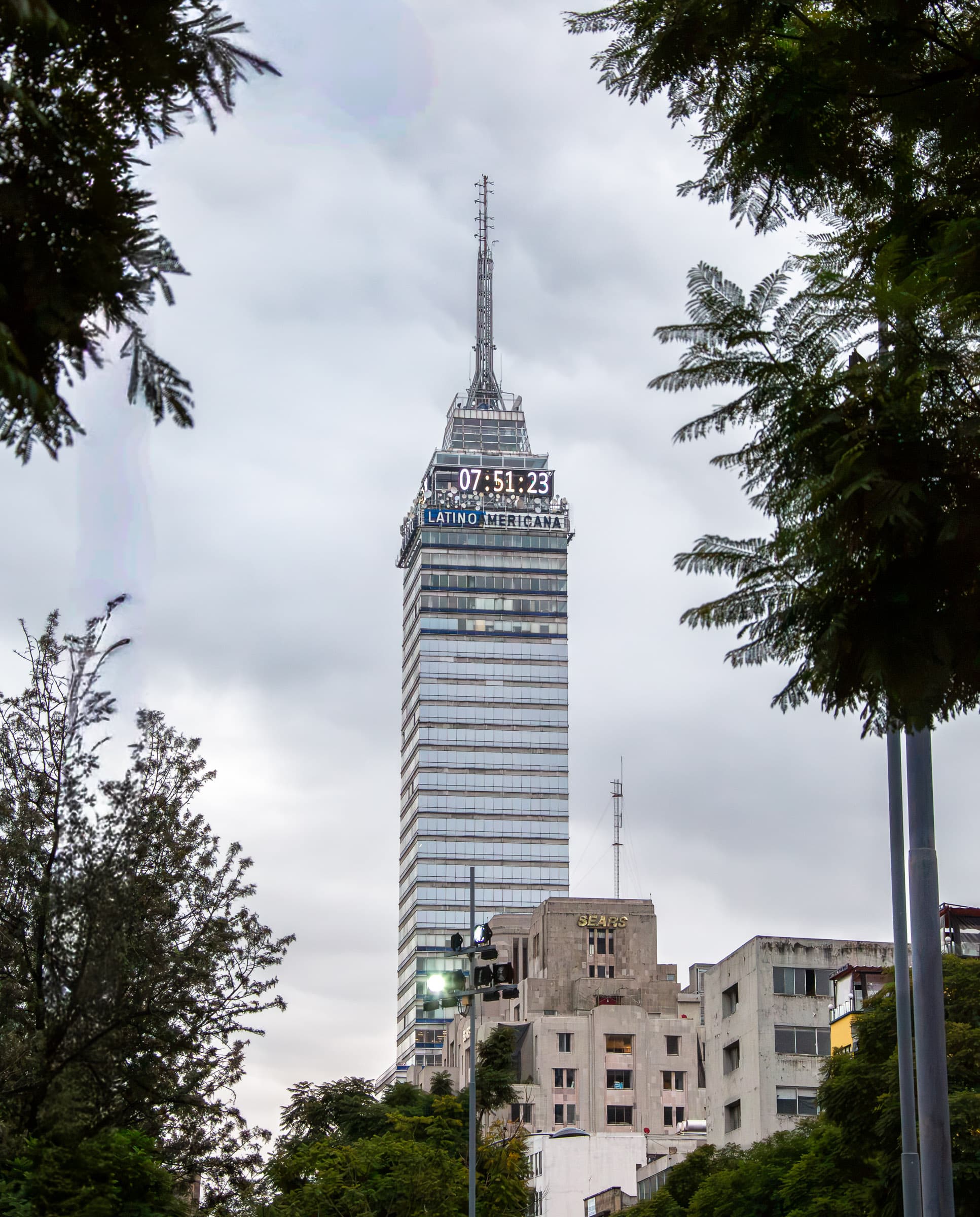 Torre Latinoamericana rising above Mexico City and framed by trees, with its illuminated clock reading 07:51:23, photographed by Francisco Colón.
