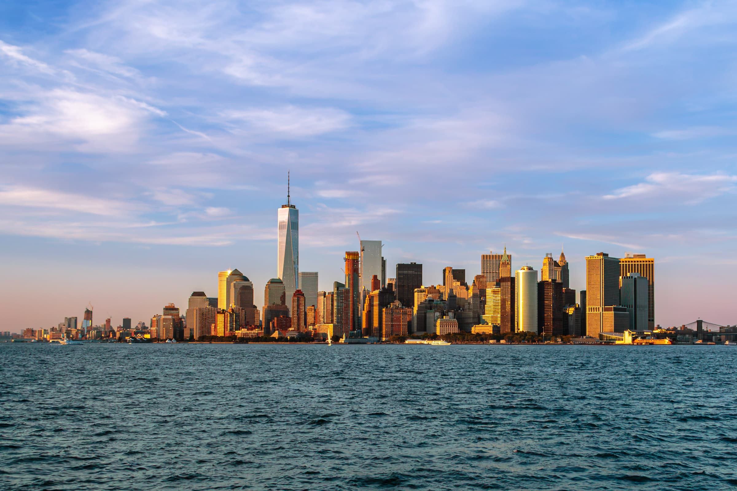 Lower Manhattan skyline and One World Trade Center illuminated by late-day light across New York Harbor beneath an expansive blue and cloud-filled sky, photographed by Francisco Colón.