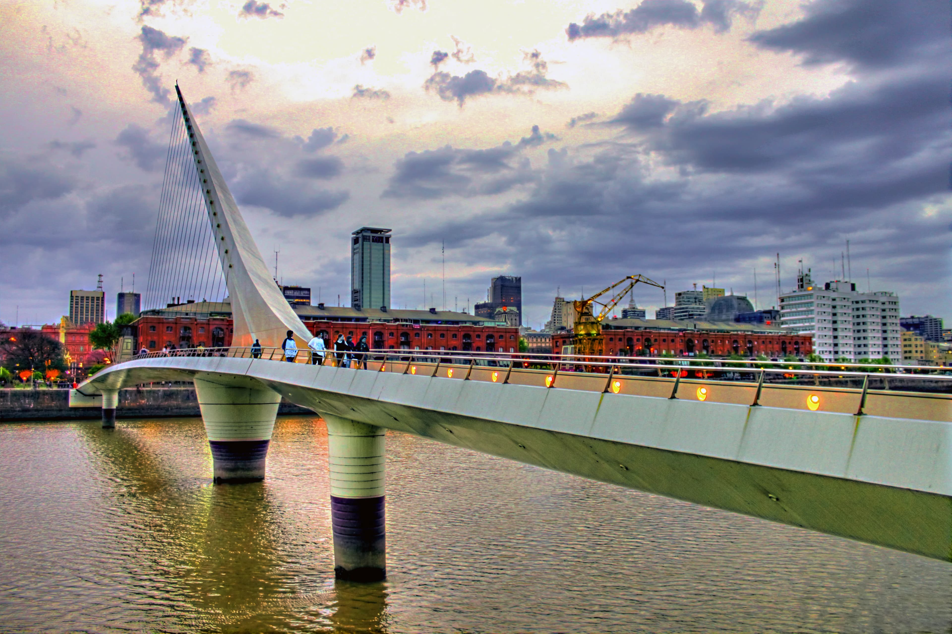 Puente de la Mujer pedestrian bridge extending across the water toward the Puerto Madero skyline beneath dramatic clouds, titled Crossing Horizons by Francisco Colón.