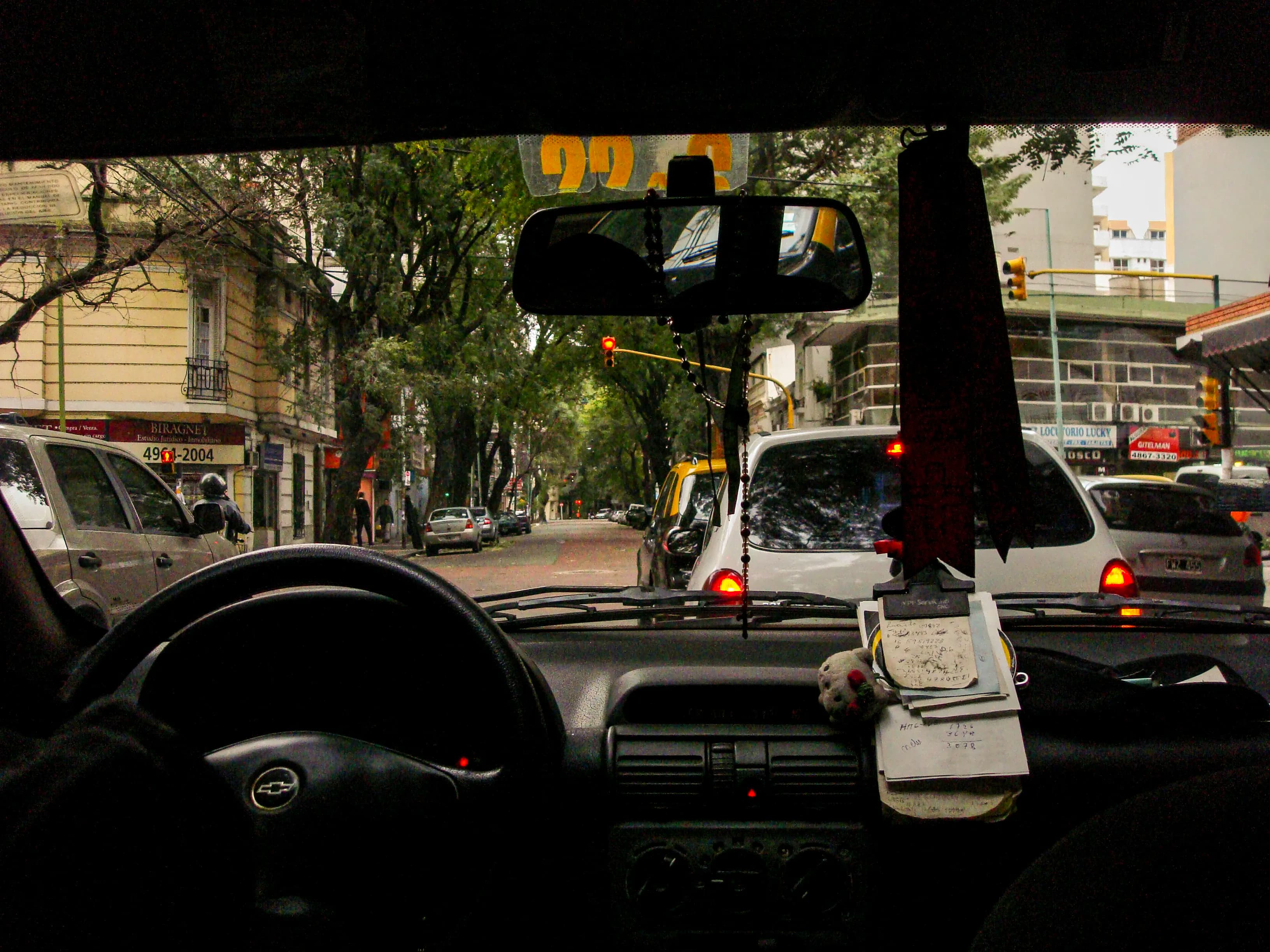 Passenger view through a taxi windshield at a red light in Buenos Aires, with dashboard objects and city buildings layered together, titled Waiting for Green by Francisco Colón.