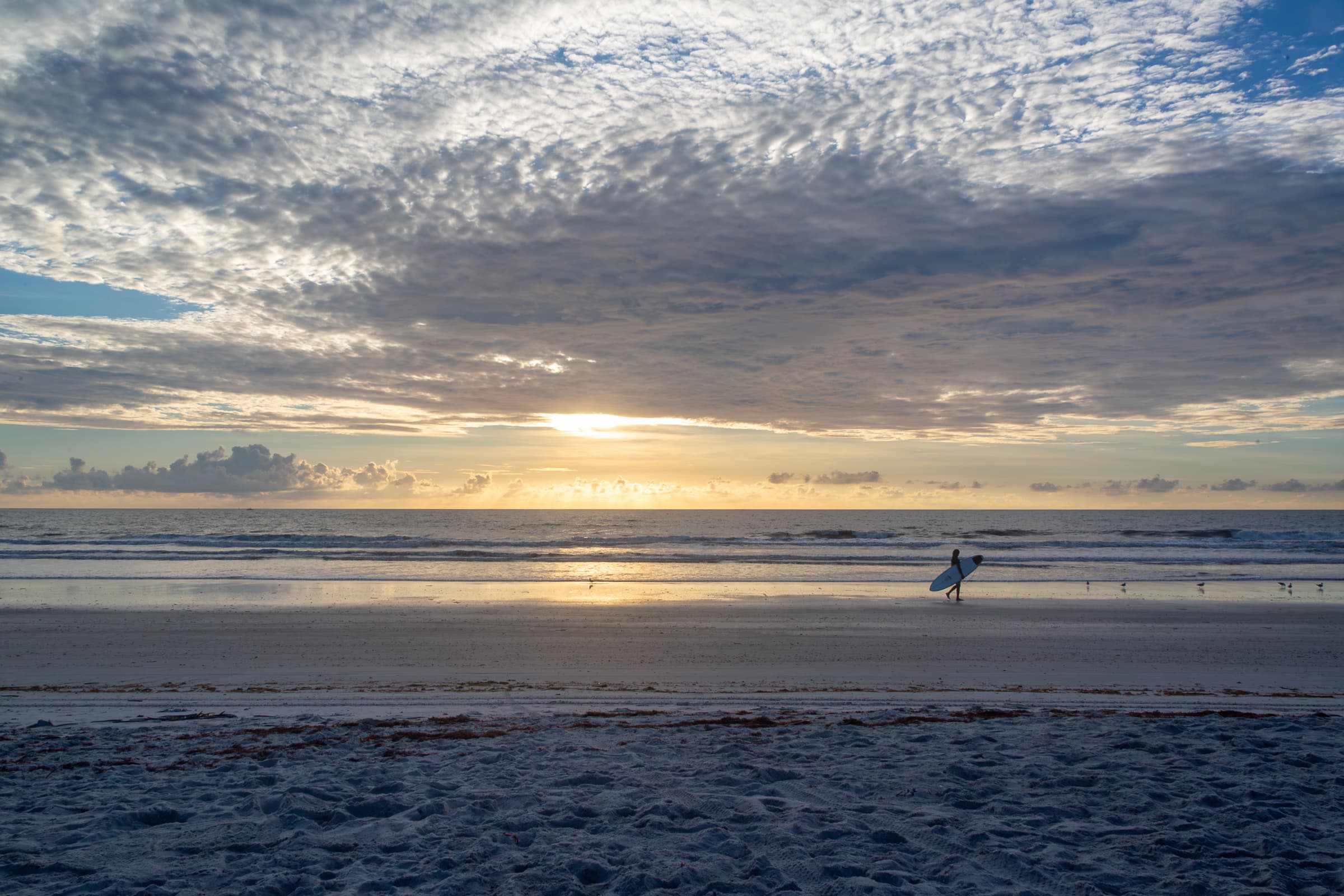 A lone surfer carrying a surfboard walks along the shoreline beneath a vast cloud-filled morning sky at Cape Canaveral, Florida, in For As Long As It Carries Me by Francisco Colón.