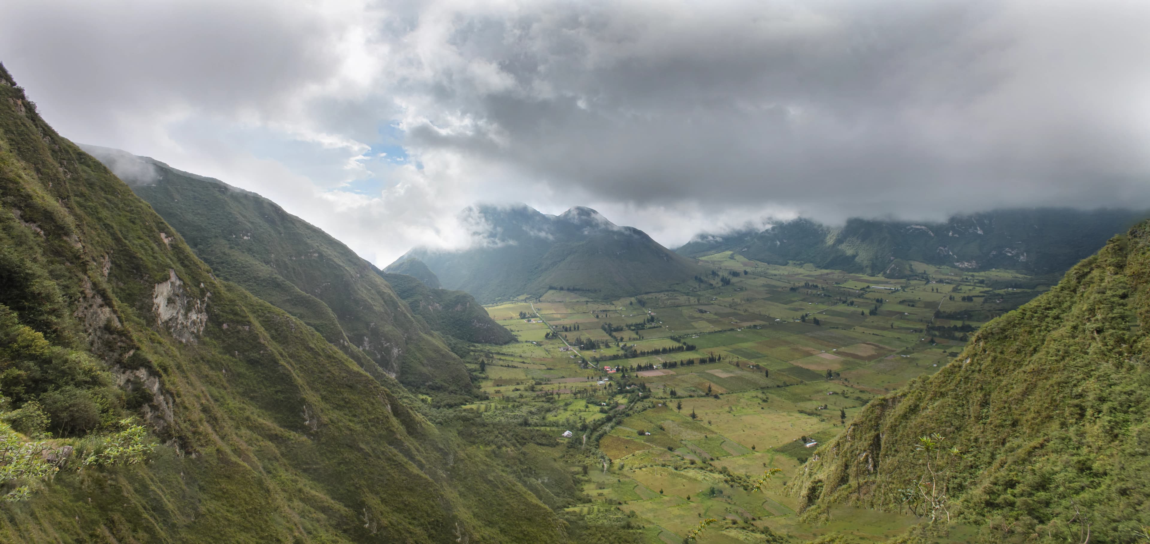 Panoramic view across the inhabited Pululahua volcanic caldera in Ecuador, with steep green crater walls, low clouds, mist, farmland, roads, and homes across the valley floor, titled Life Inside the Caldera by Francisco Colón.