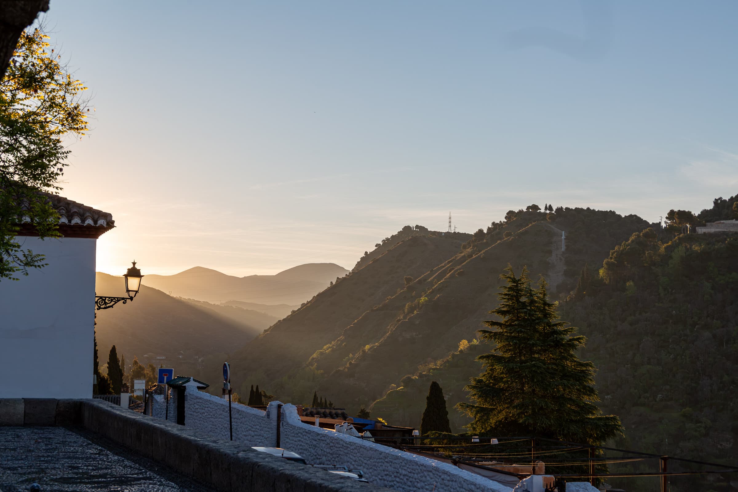 Morning sunlight reaches across layered mountain ridges and a valley near Granada, Spain, with a shadowed streetlamp, whitewashed buildings, and an evergreen tree in the foreground.