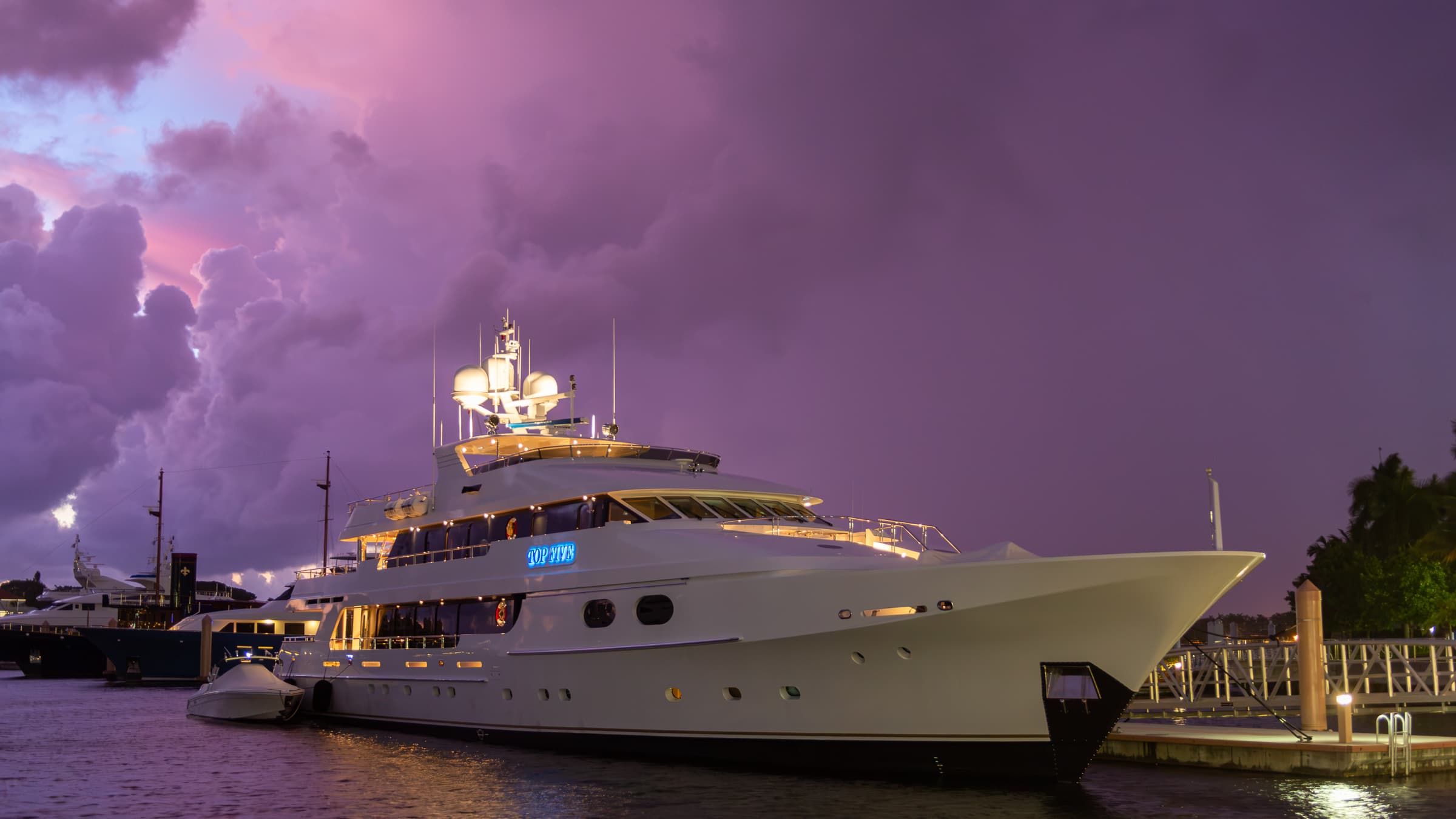 A large illuminated yacht moored at the West Palm Beach waterfront beneath expansive violet and pink clouds during the early morning.