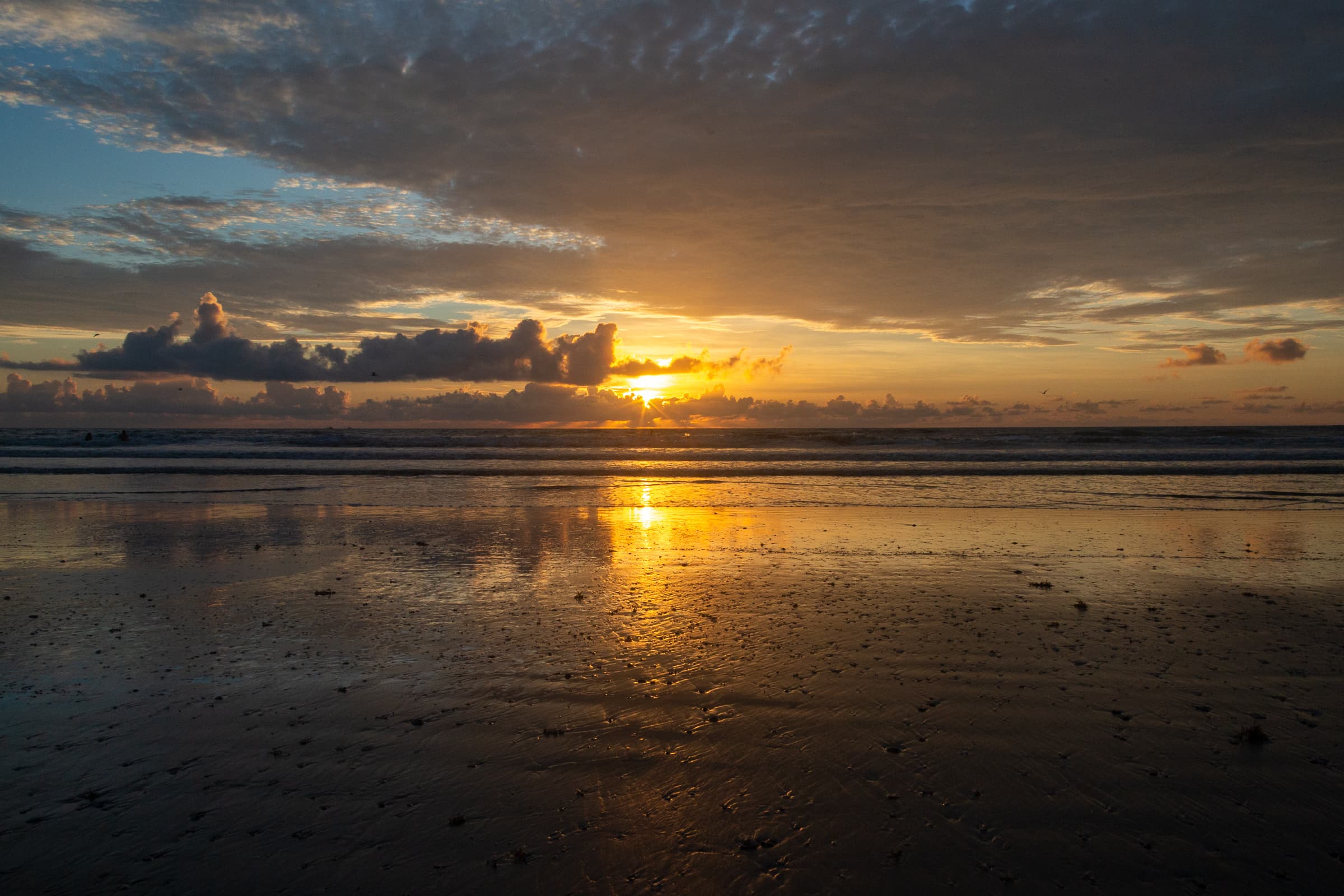 Sunrise over the Atlantic Ocean with dark wet sand in the foreground, reflected sunlight across the beach and water, and a large cloud layer above the illuminated horizon.