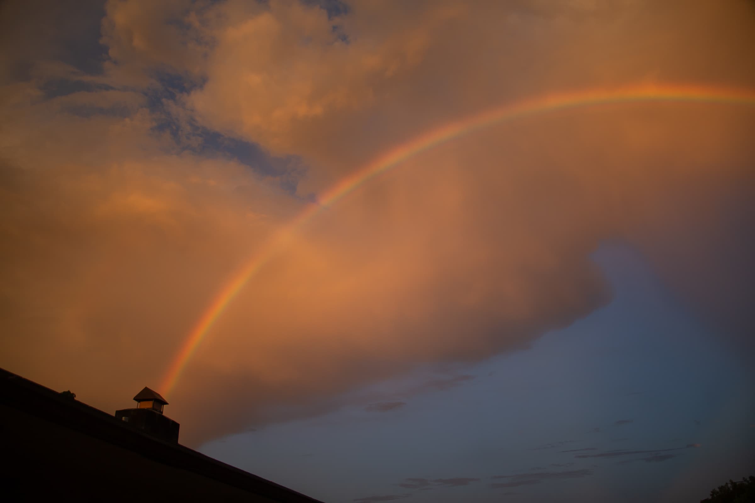 Rainbow appearing to rise from the chimney of Francisco Colón's home beneath warm clouds and blue evening sky.