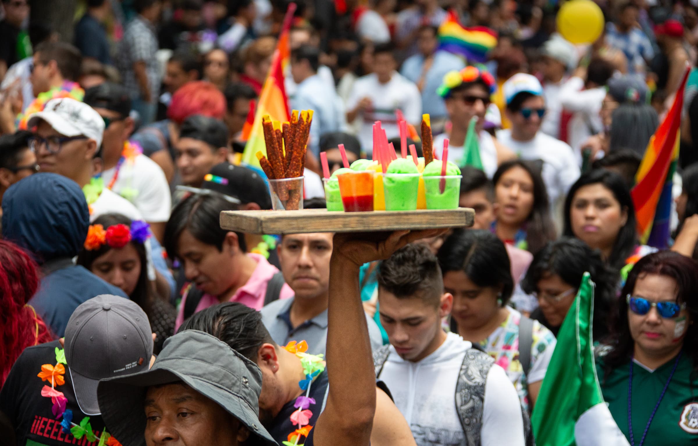 A partially concealed street vendor raises a wooden tray of colorful refreshments and snacks above a dense Pride march crowd in Mexico City.