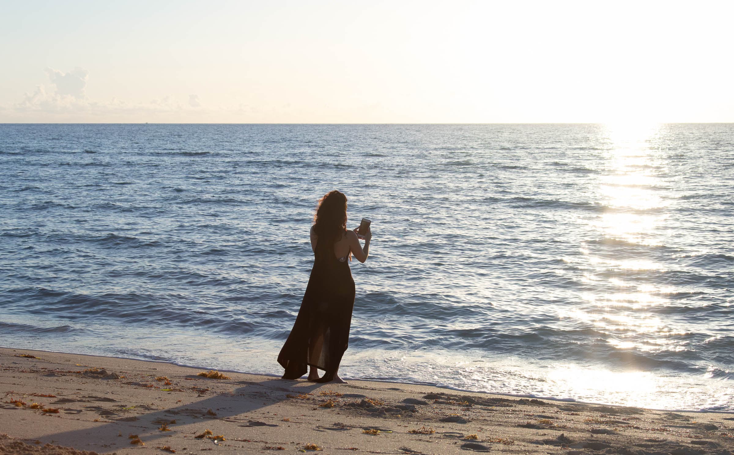 Melina Almodóvar stands in silhouette at the Miami Beach shoreline, photographing the sunrise as her long shadow stretches across the sand.