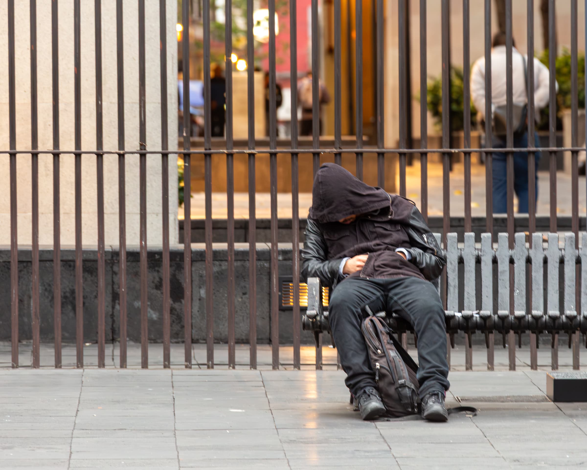 A hooded man rests on a public bench with a backpack between his legs during the early morning in Mexico City, while people move behind an iron fence.