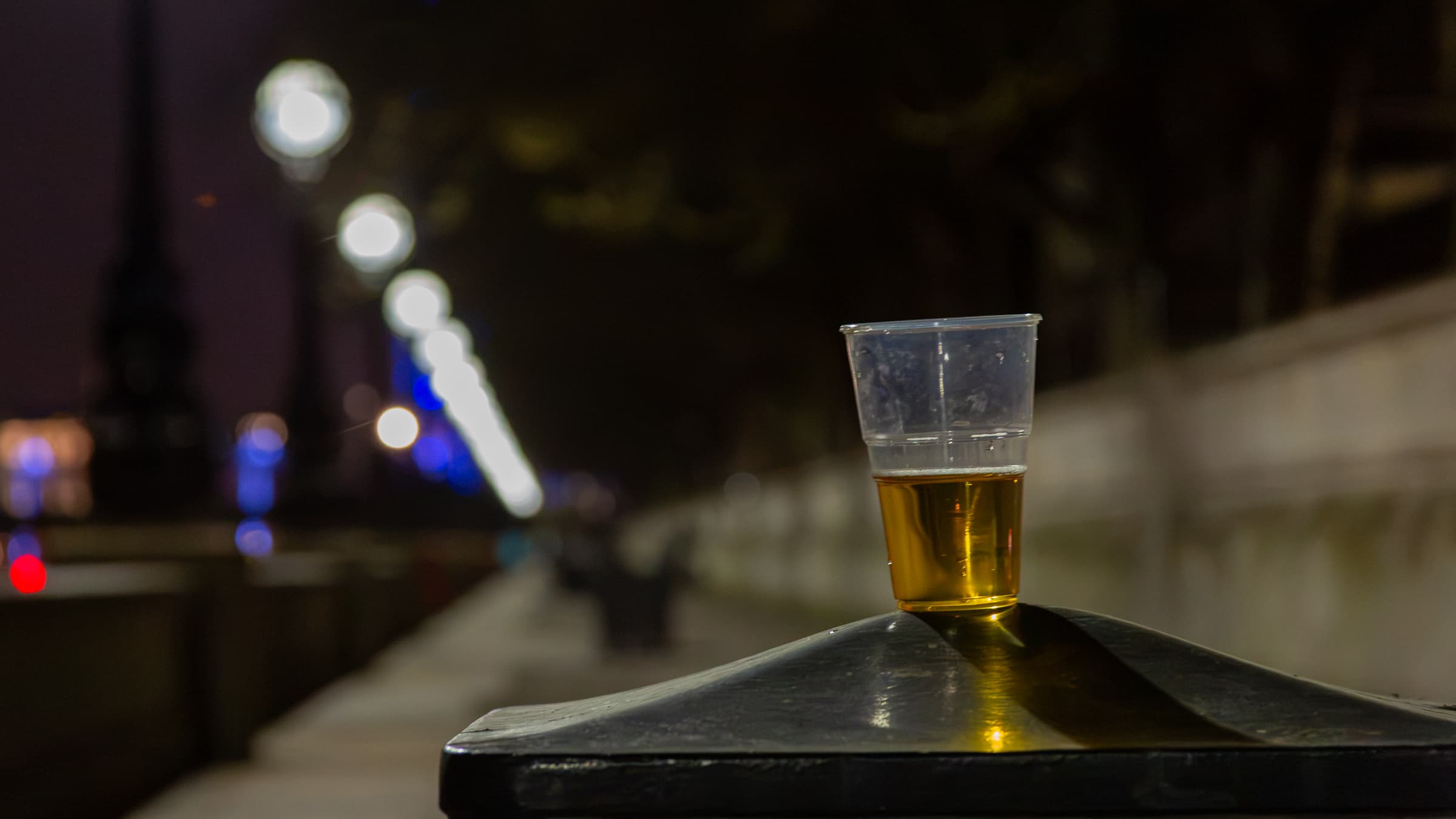 A clear plastic cup partly filled with amber beer rests on a dark metal surface beside the Thames, with softly blurred streetlights receding behind it.