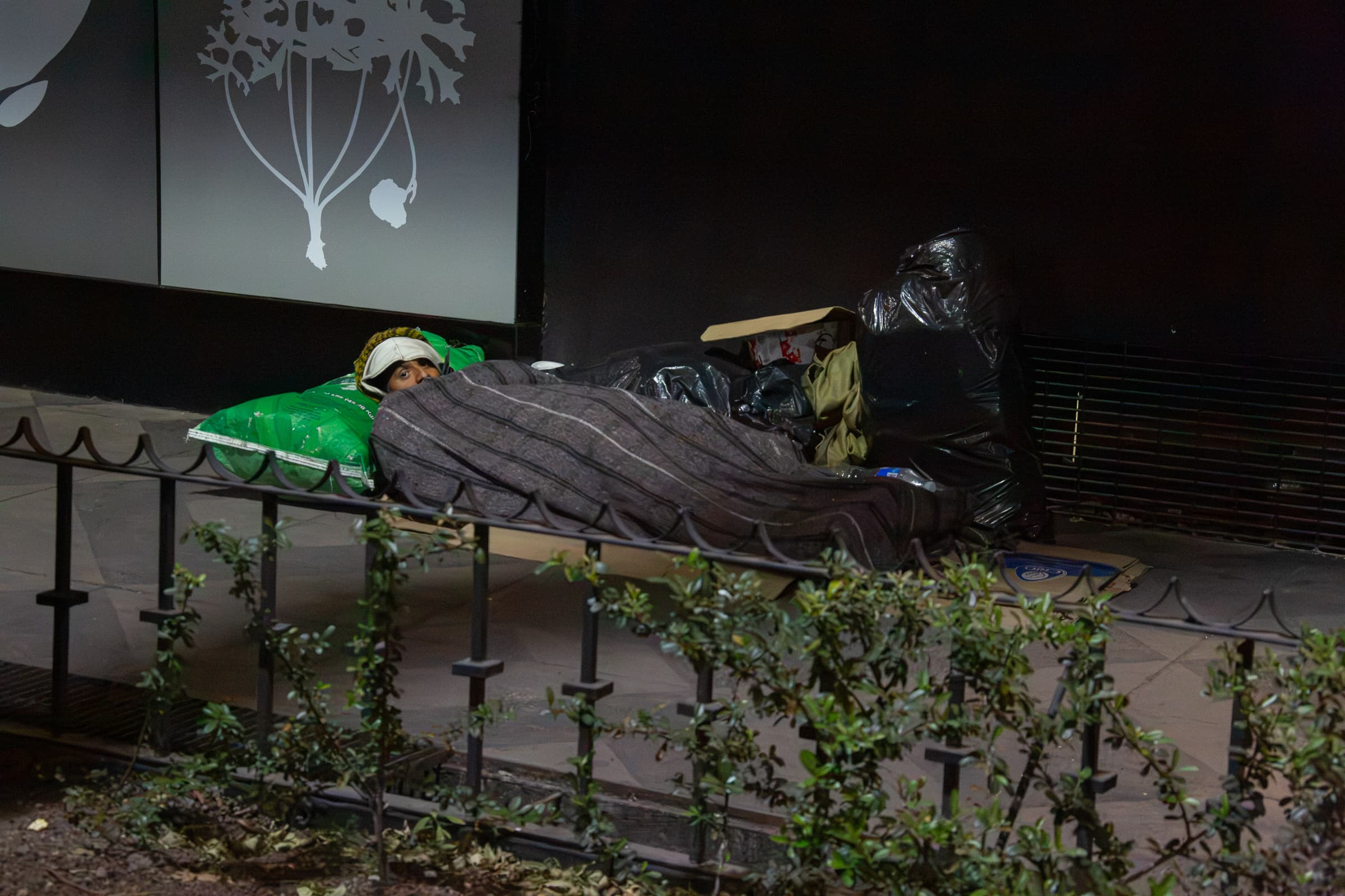 An unidentified man lying beneath a blanket beside his belongings in Mexico City at night, looking briefly toward the photographer from behind a foreground railing and vegetation.
