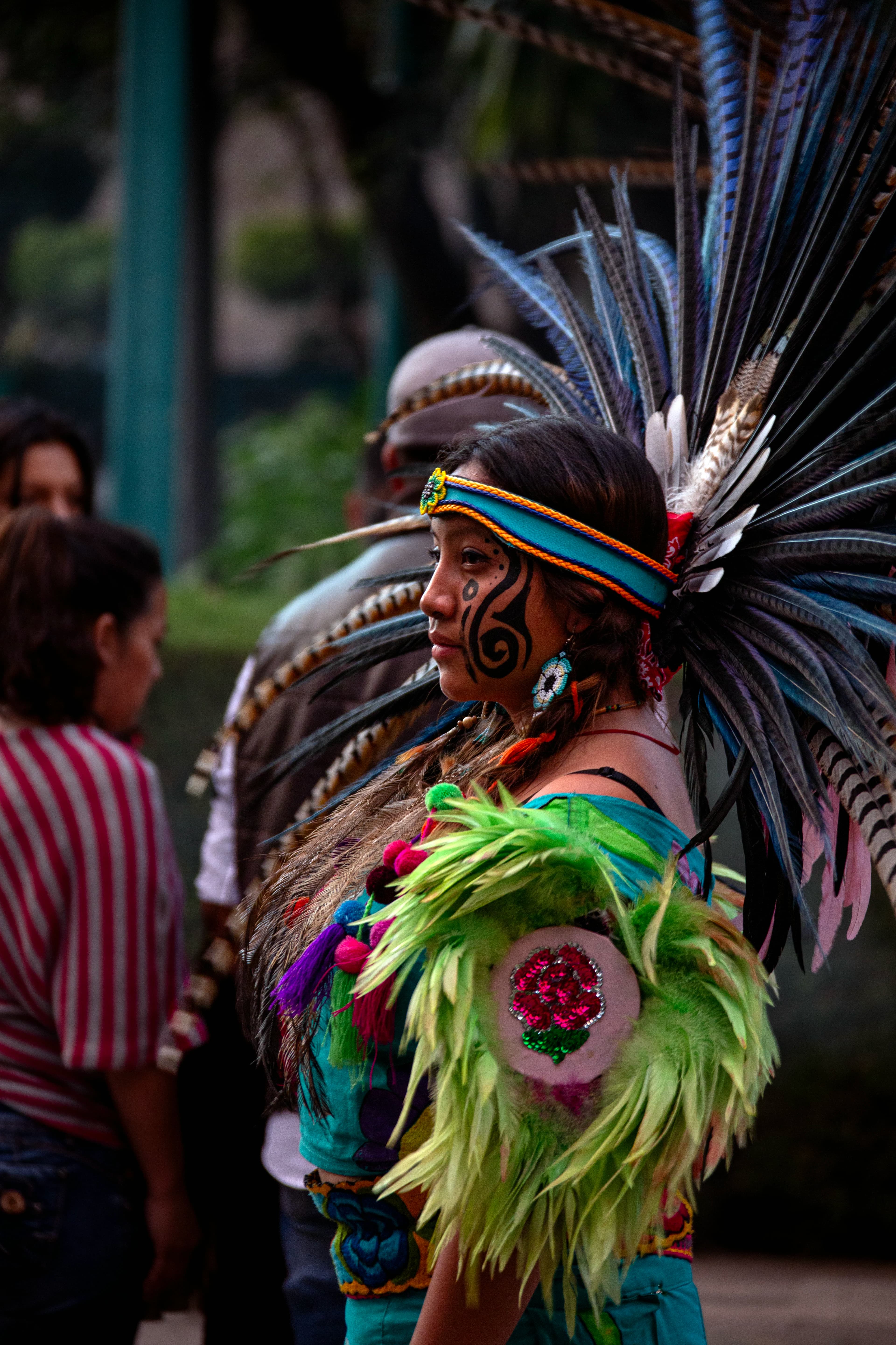 Woman photographed in profile at a cultural gathering in Mexico City, wearing elaborate feathered regalia and facial decoration while people gather softly out of focus behind her.