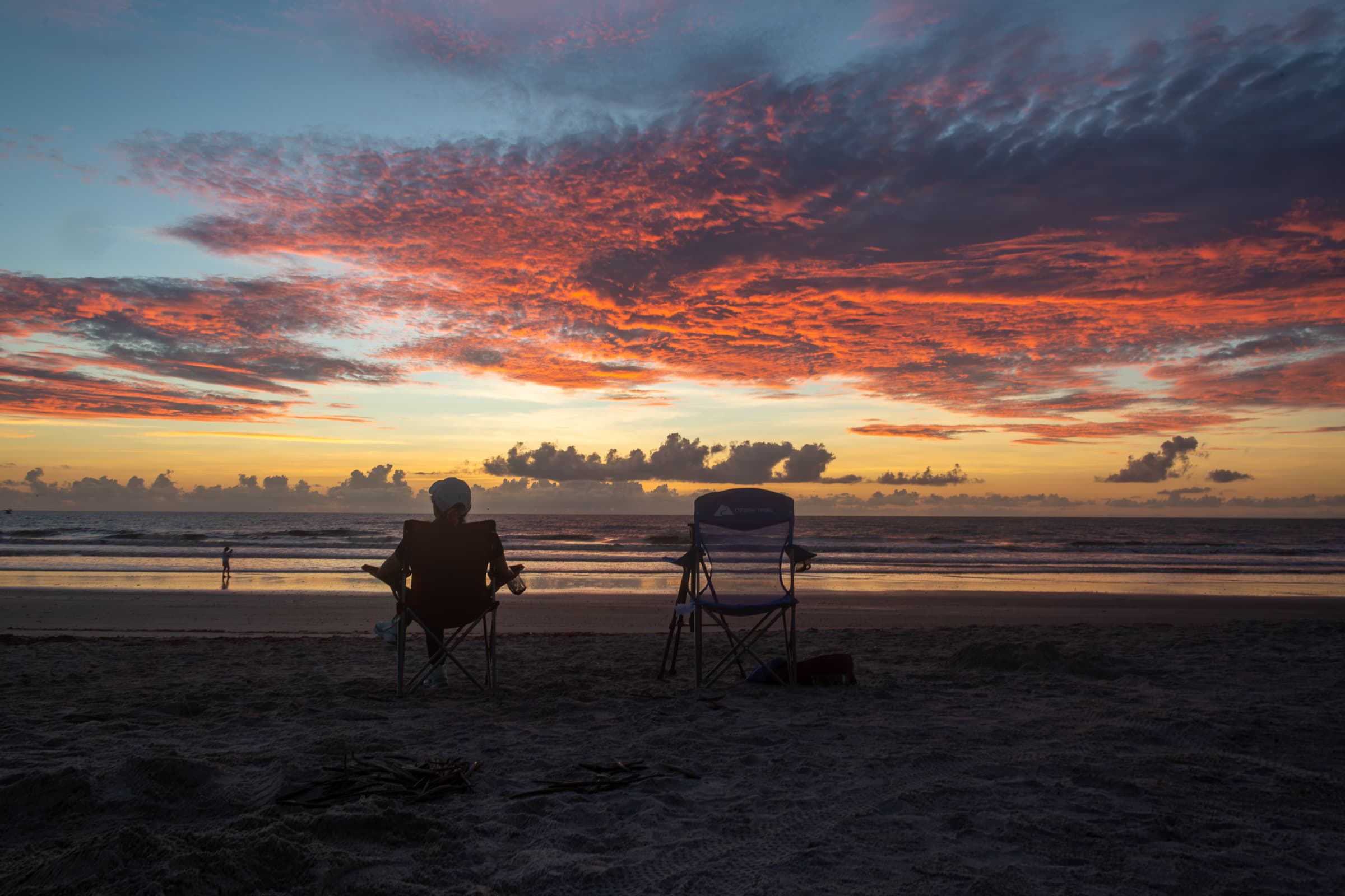 A seated person and an empty beach chair face the Atlantic beneath a dramatic red and orange sunrise at Cape Canaveral, Florida, in The Chair Beside Her by Francisco Colón.