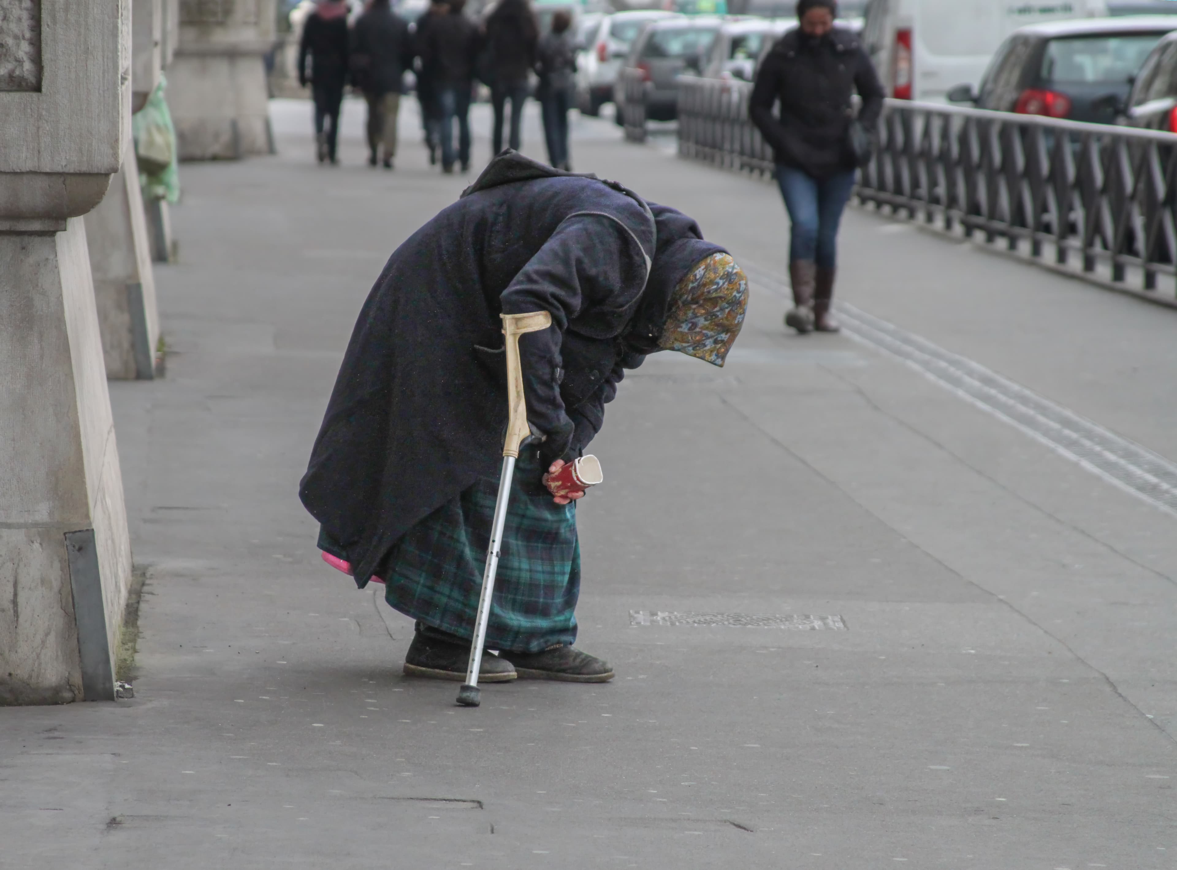 An unidentified person bends forward over a crutch while holding a small cup as pedestrians continue along a sidewalk in Paris.