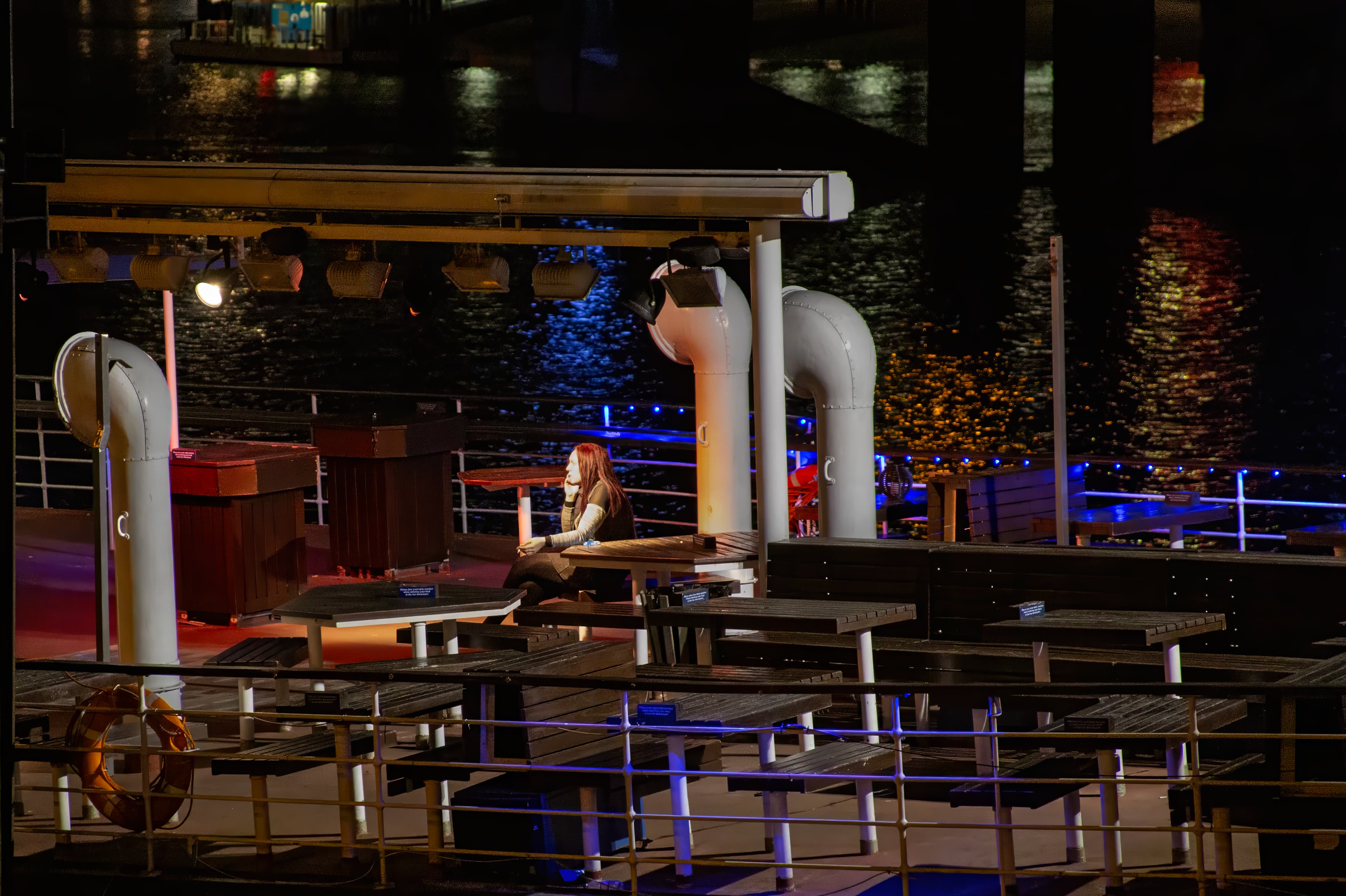 A solitary woman sits aboard a boat on the River Thames in London, surrounded by rows of empty tables and chairs beneath warm and blue nighttime lights.