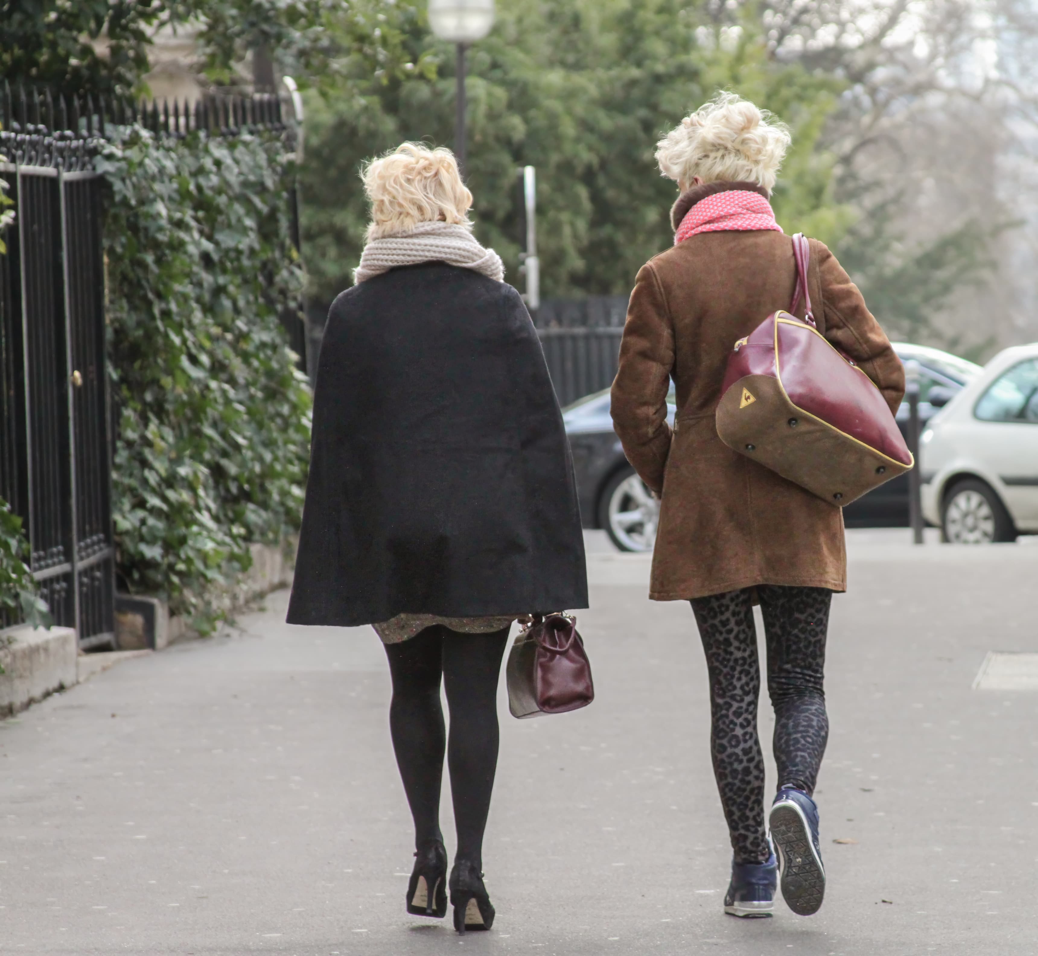 Two blonde women wearing long coats, scarves, and coordinated handbags walk together along a sidewalk in Paris.