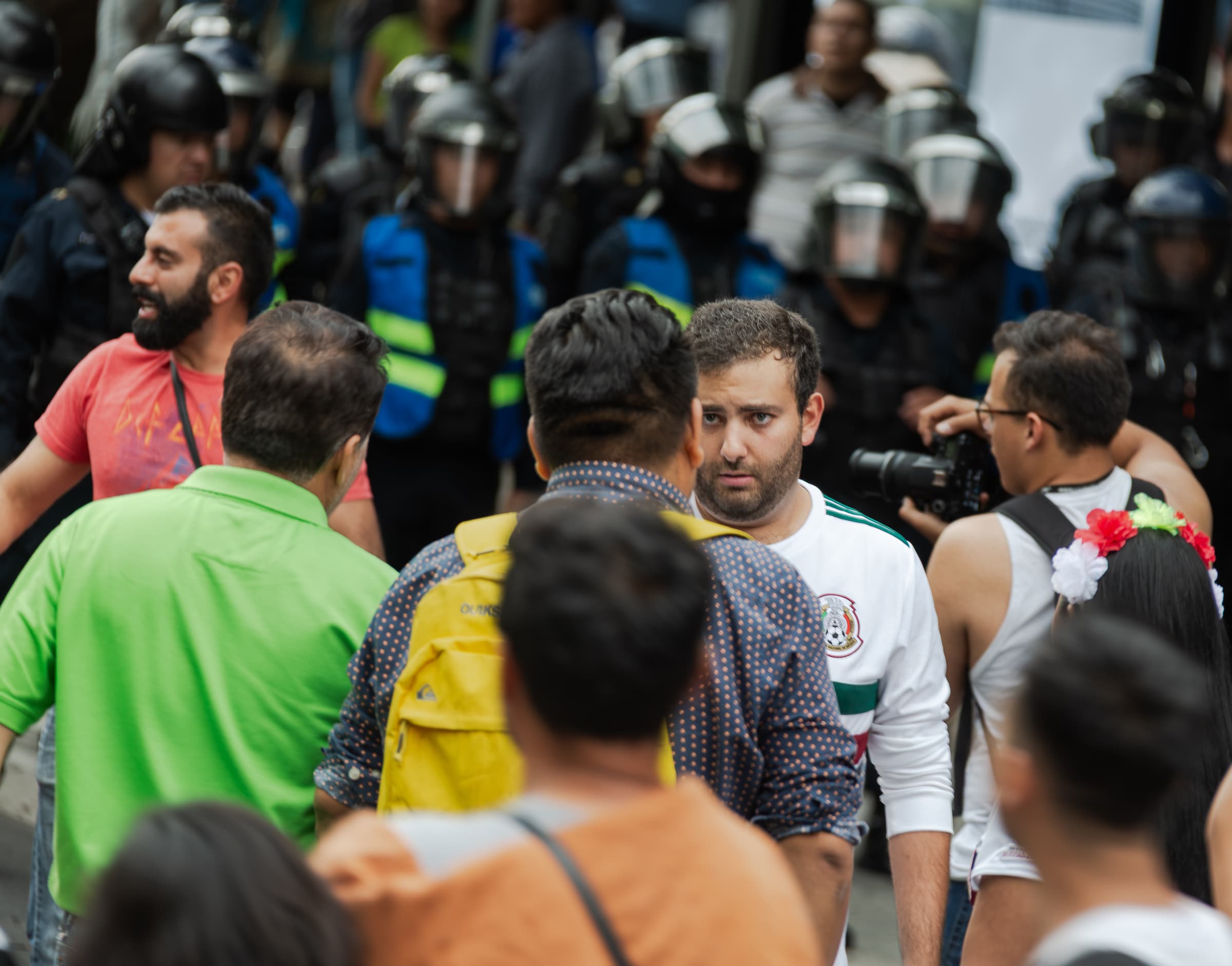 A focused man in a white Mexico football shirt listens within a crowded animal-rights demonstration as a line of helmeted police stands behind him in Mexico City.