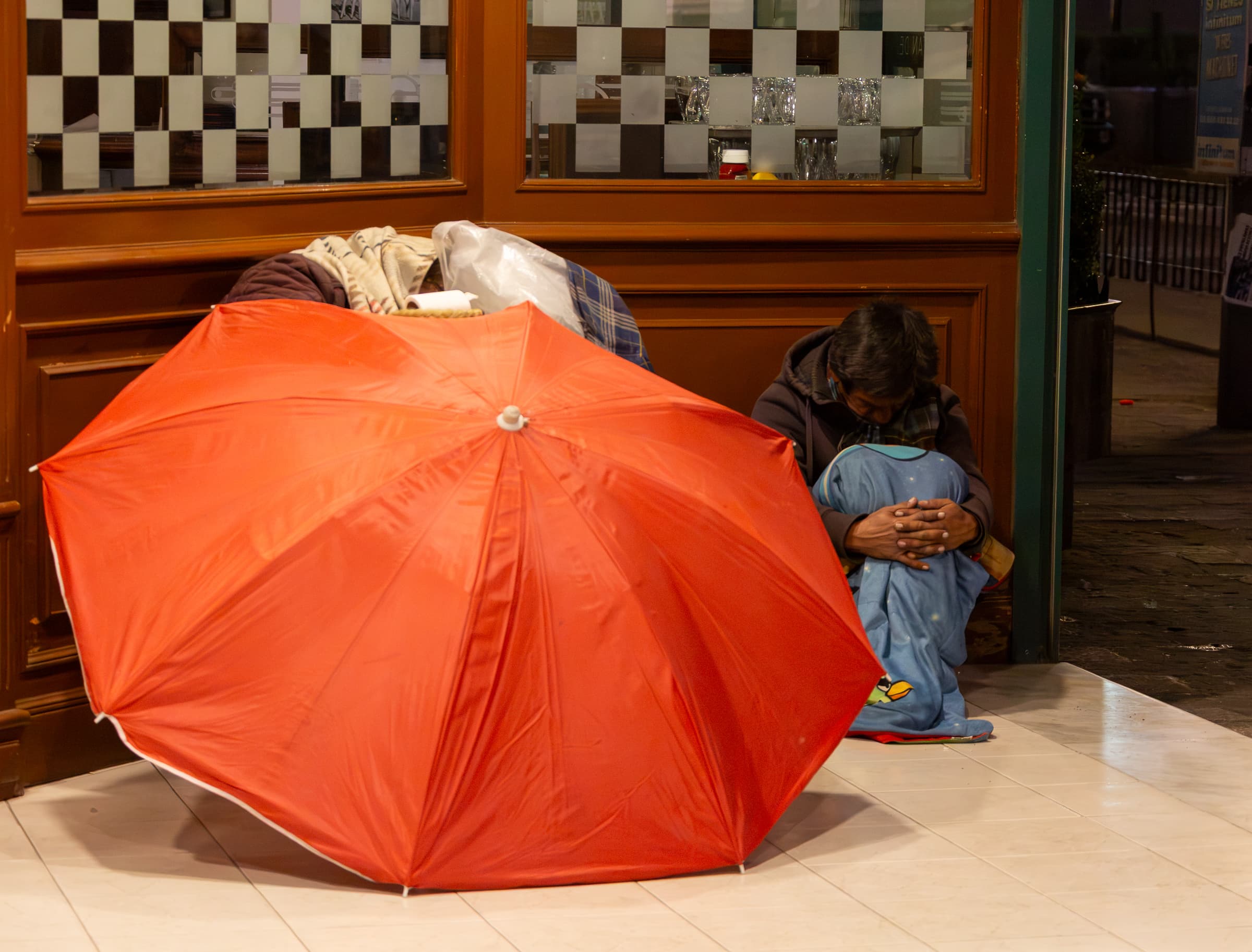 A solitary person sits curled inward beside a storefront in Mexico City while a large red umbrella fills the foreground.