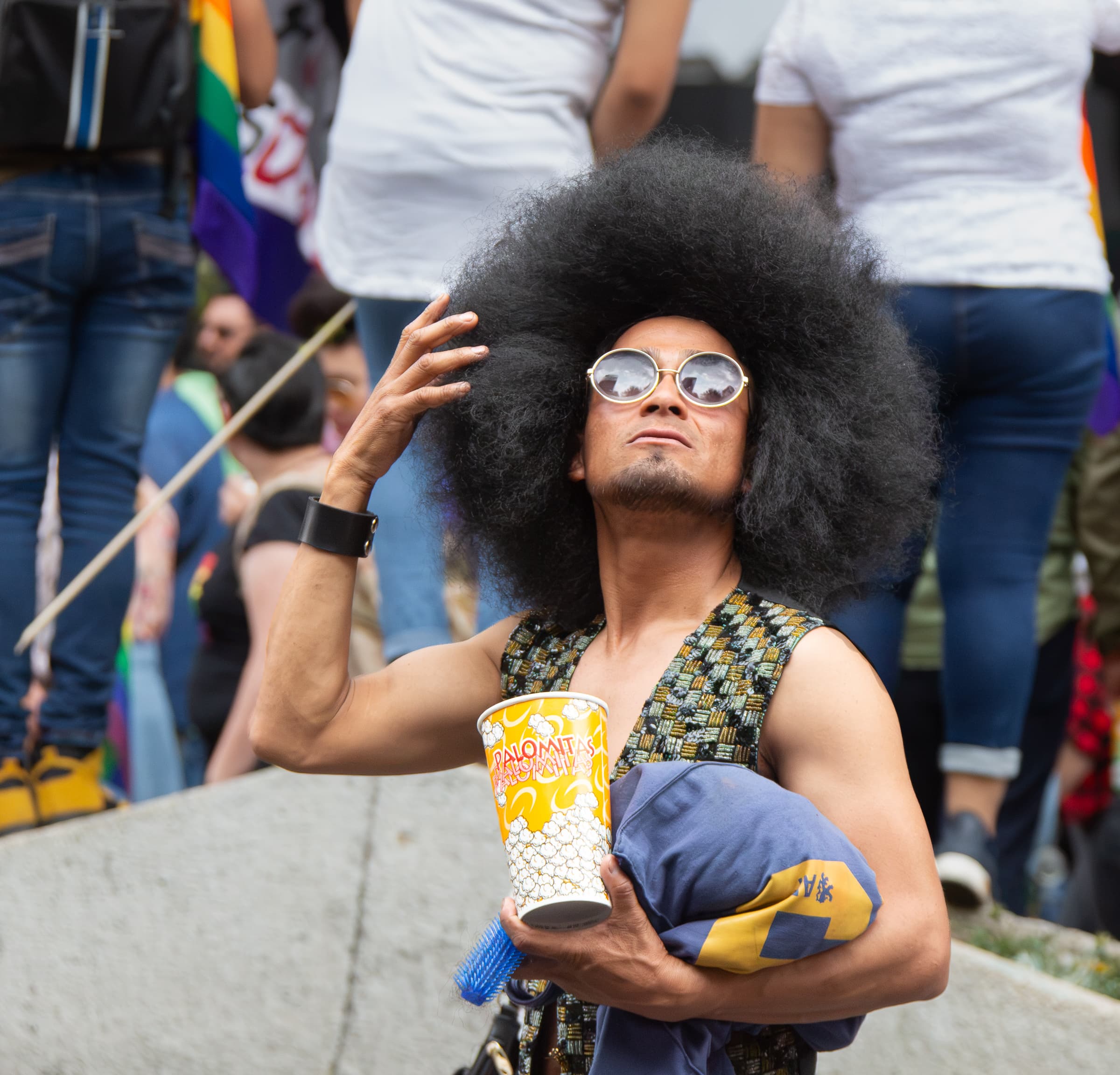 A man with expansive dark hair and round reflective glasses raises one hand while holding folded clothing and a popcorn cup during the Mexico City Pride march.