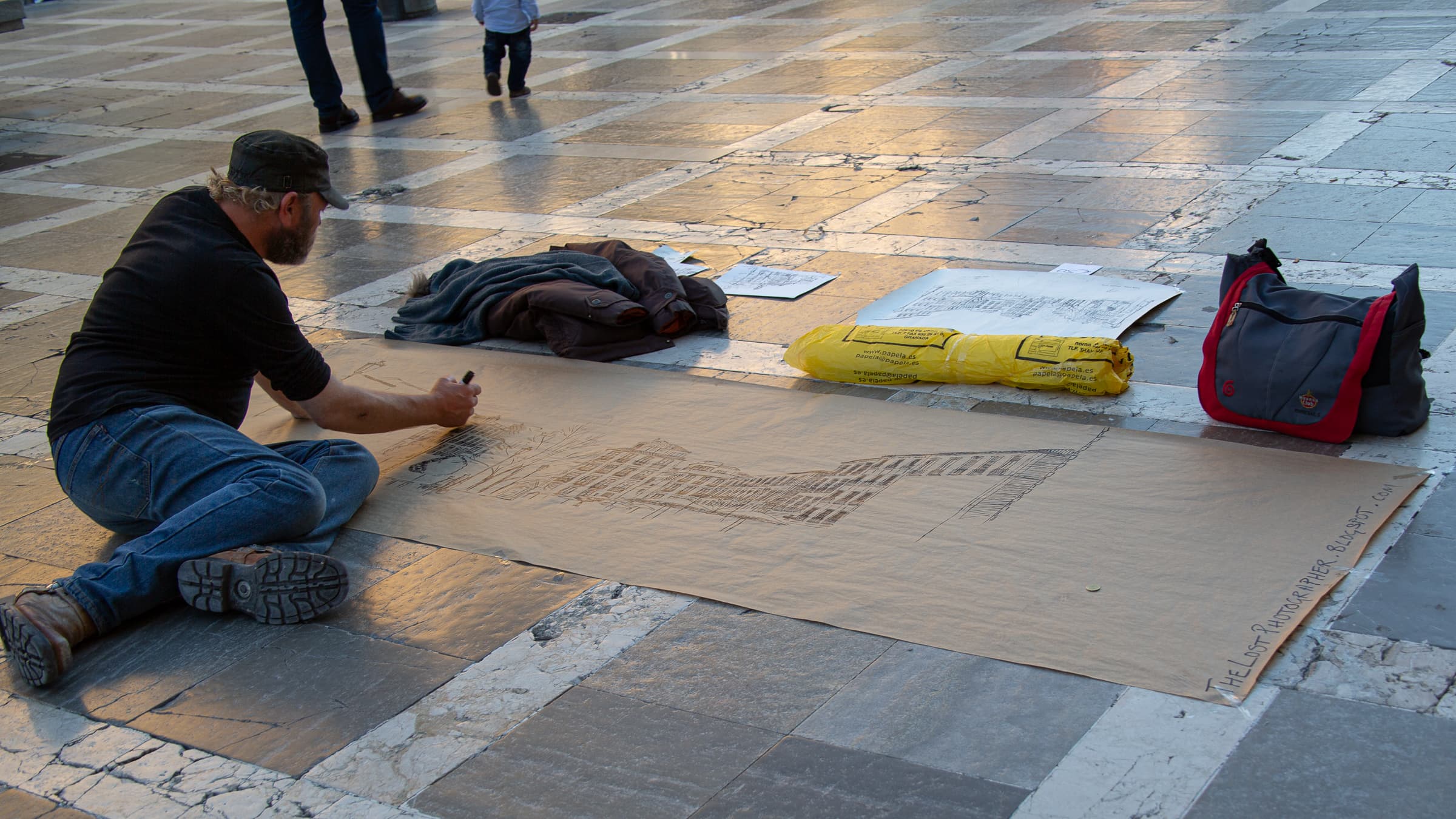 Artist John Haydn Colley seated on the pavement in Granada, Spain, creating a large architectural drawing on cardboard while pedestrians pass behind him.