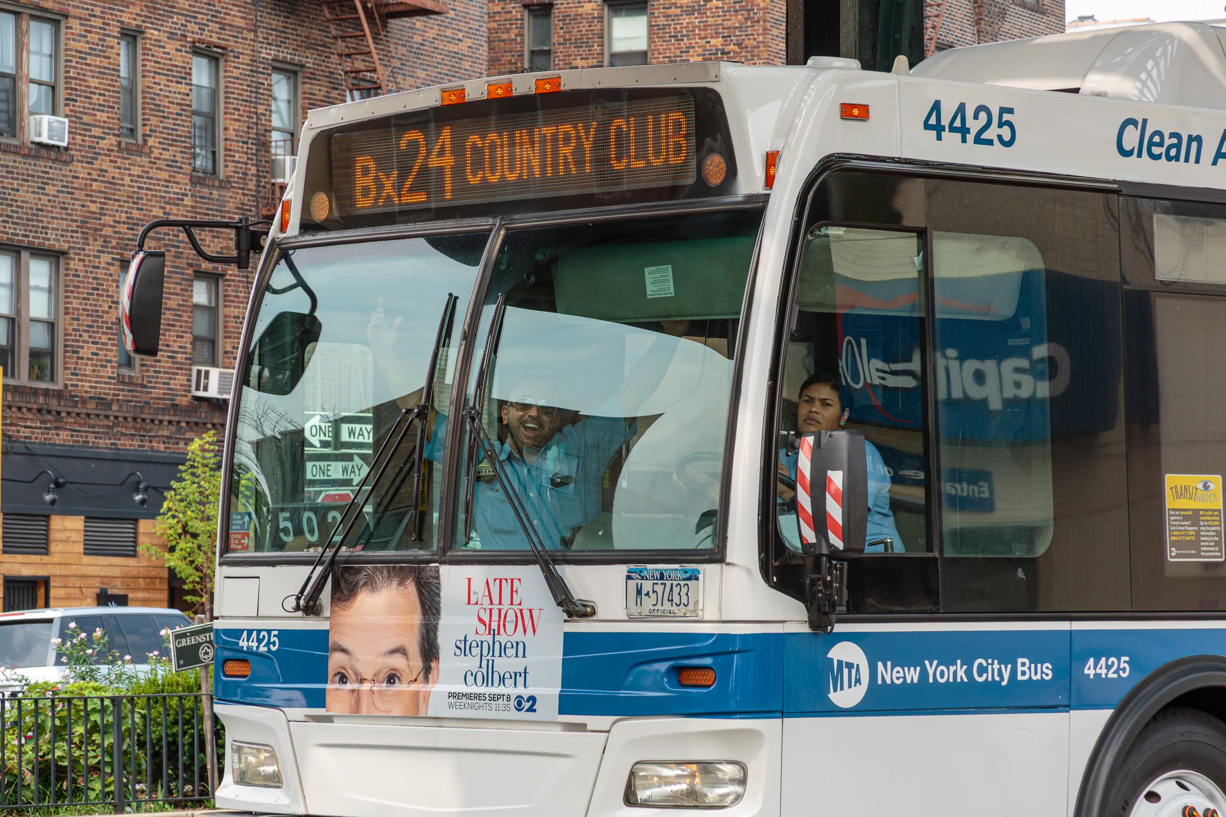 New York City Bx24 bus number 4425 with a smiling driver looking toward the photographer and a Stephen Colbert advertisement on the front, photographed by Francisco Colón.