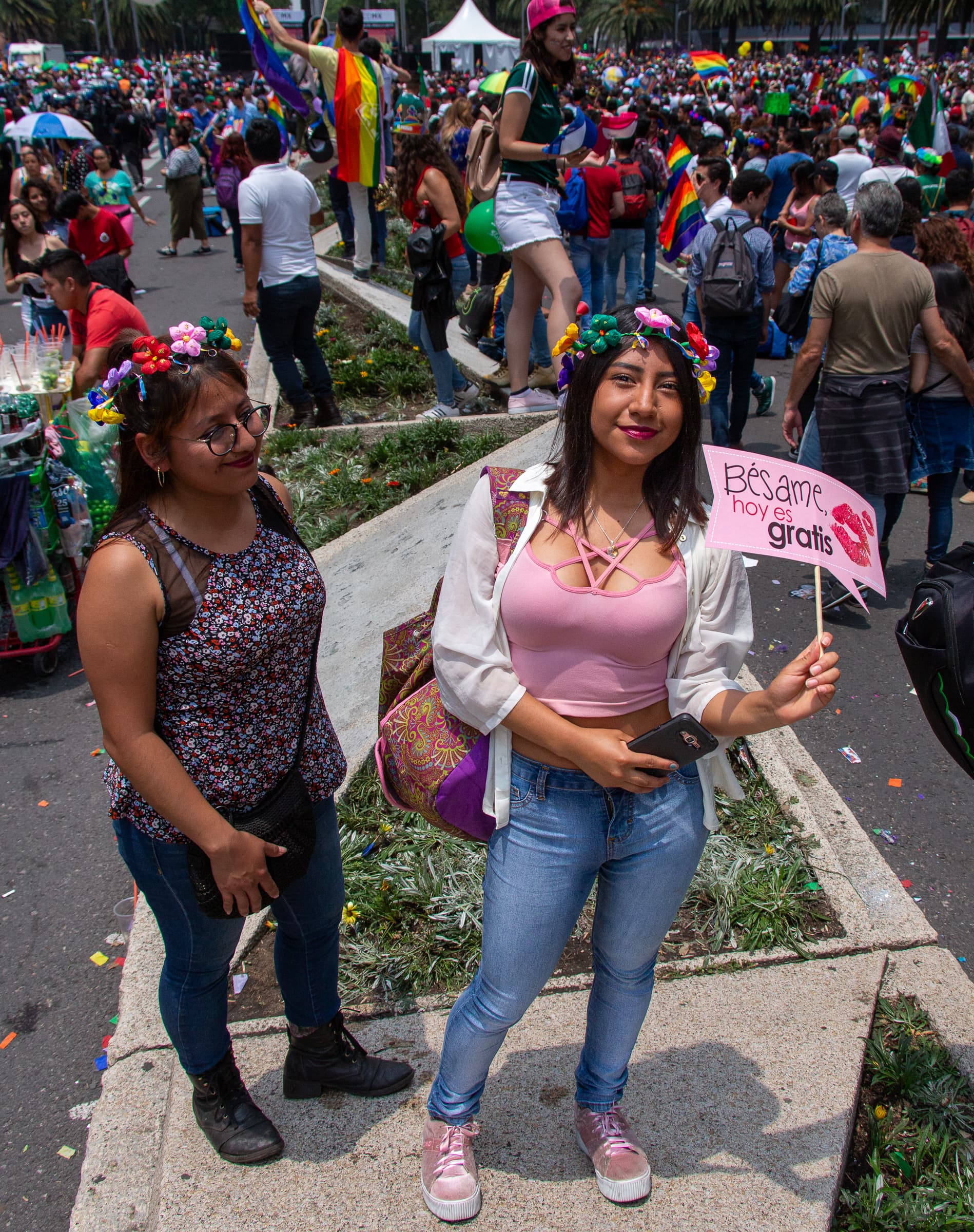 A smiling woman at the Mexico City Pride march holds a handwritten sign reading Bésame, hoy es gratis while another woman and a colorful crowd stand behind her.