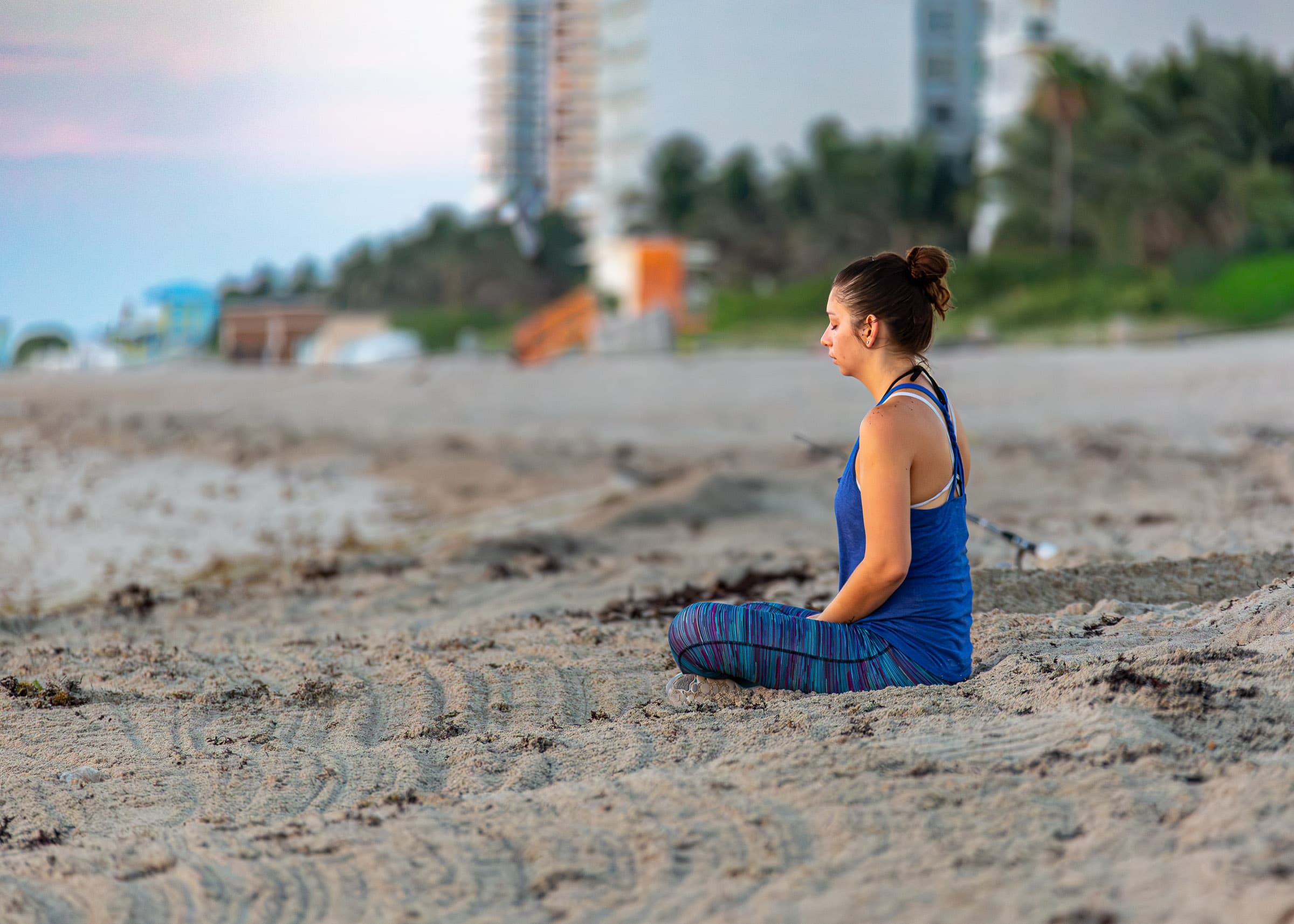 Woman meditating alone on a Florida beach in the early morning, seated in the sand and facing toward the rising sun, photographed from a distance by Francisco Colón.