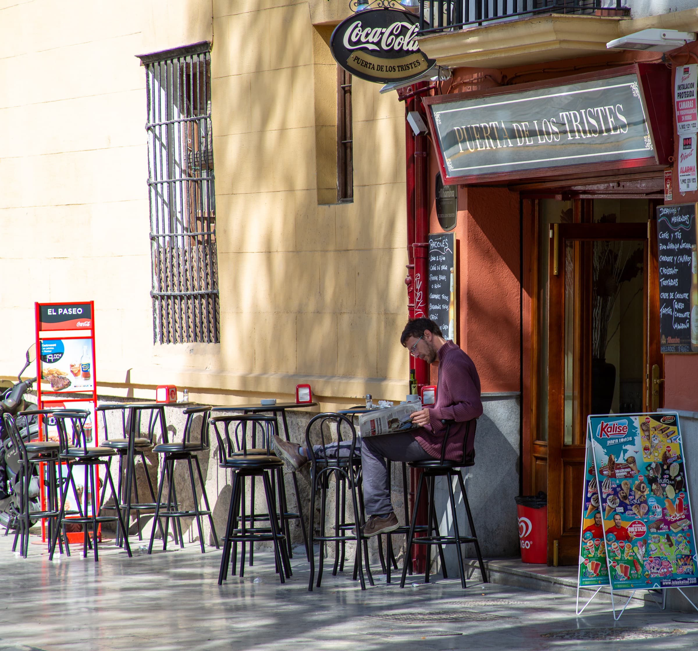 A solitary man reads a newspaper outside Puerta de los Tristes in Granada, surrounded by empty café tables and chairs in the late-morning light.