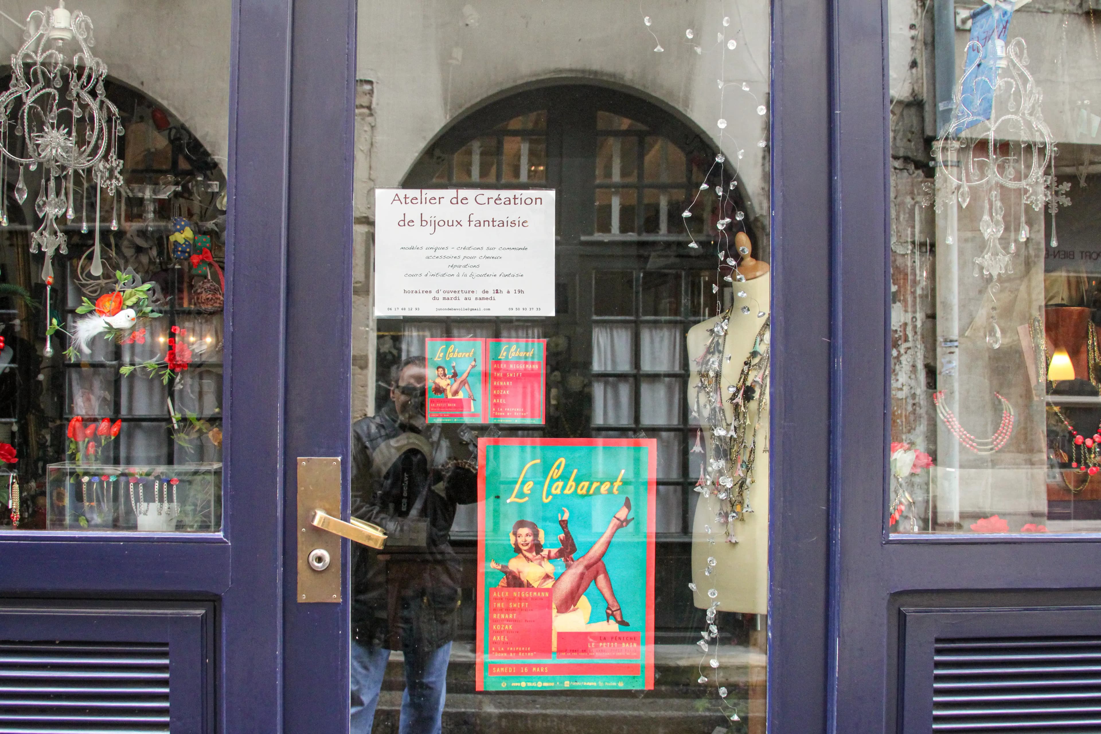 A Paris jewelry-shop window displays costume jewelry, crystal ornaments, a mannequin, and a colorful cabaret poster while the photographer appears reflected in the glass.