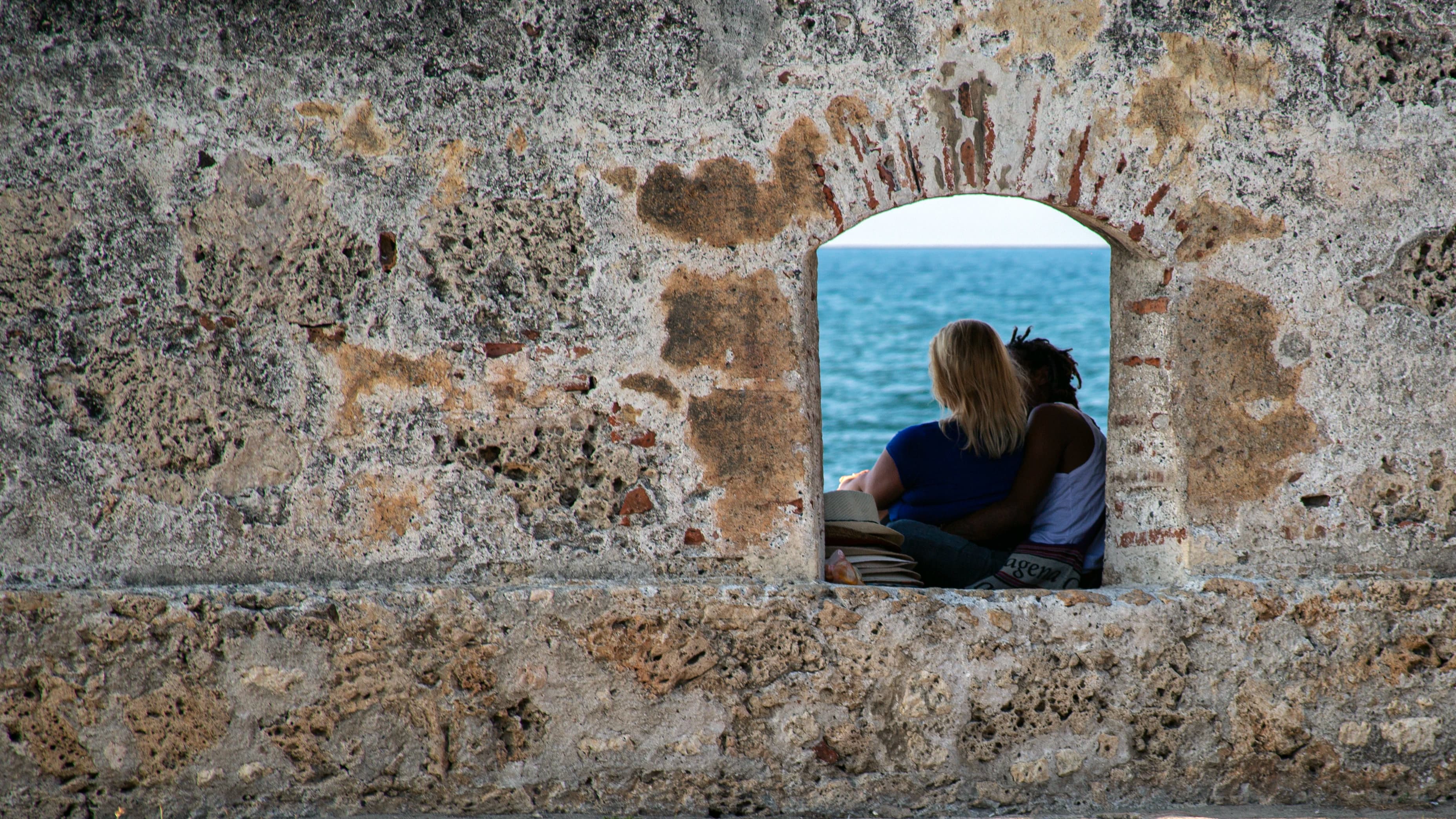 Two people sit closely together within a small arched opening in a weathered stone wall overlooking the sea in Cartagena, Colombia.