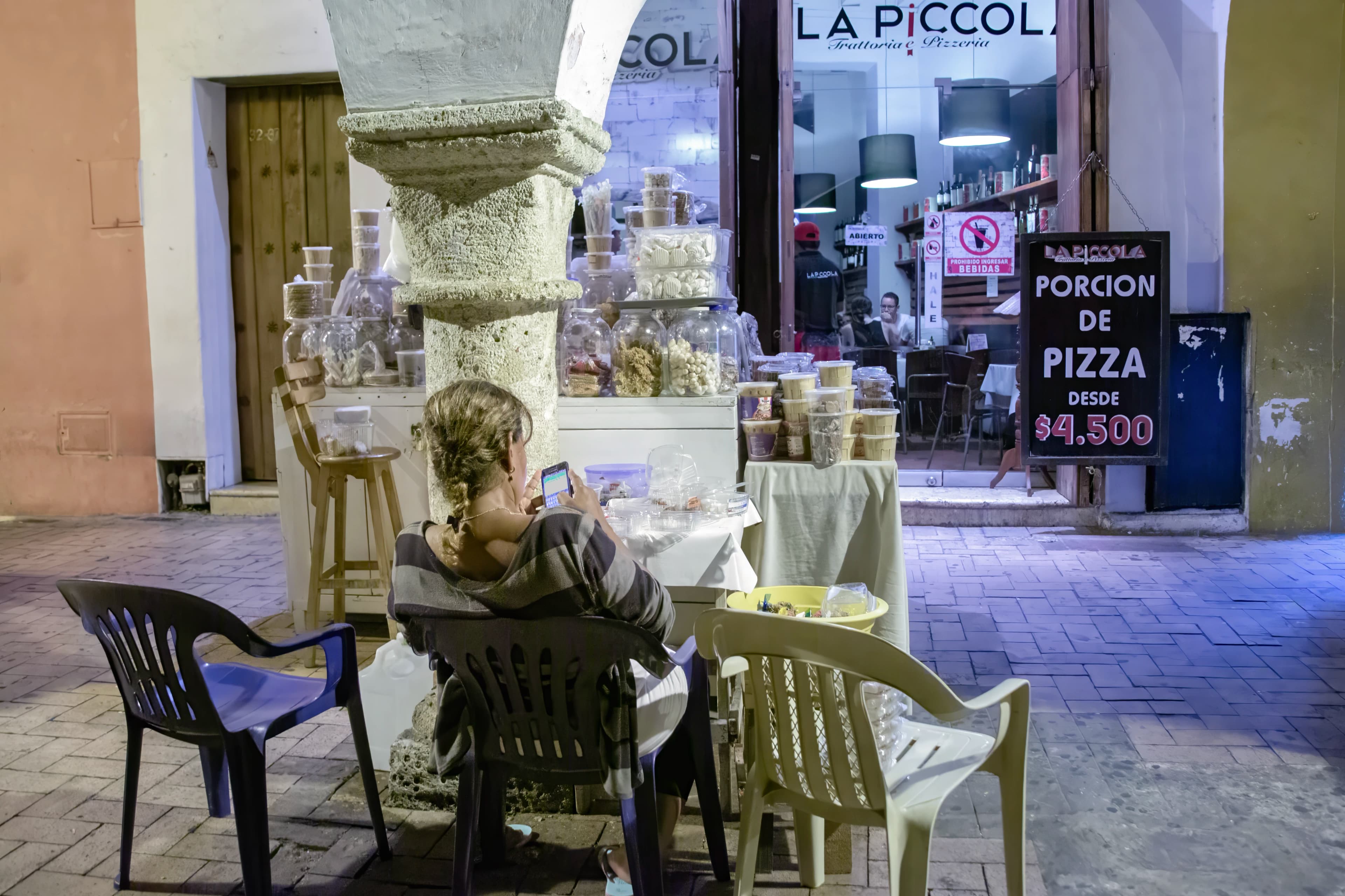 A woman sits with her back toward the camera while looking at a phone among empty chairs and a street display outside an illuminated restaurant in Cartagena, Colombia.