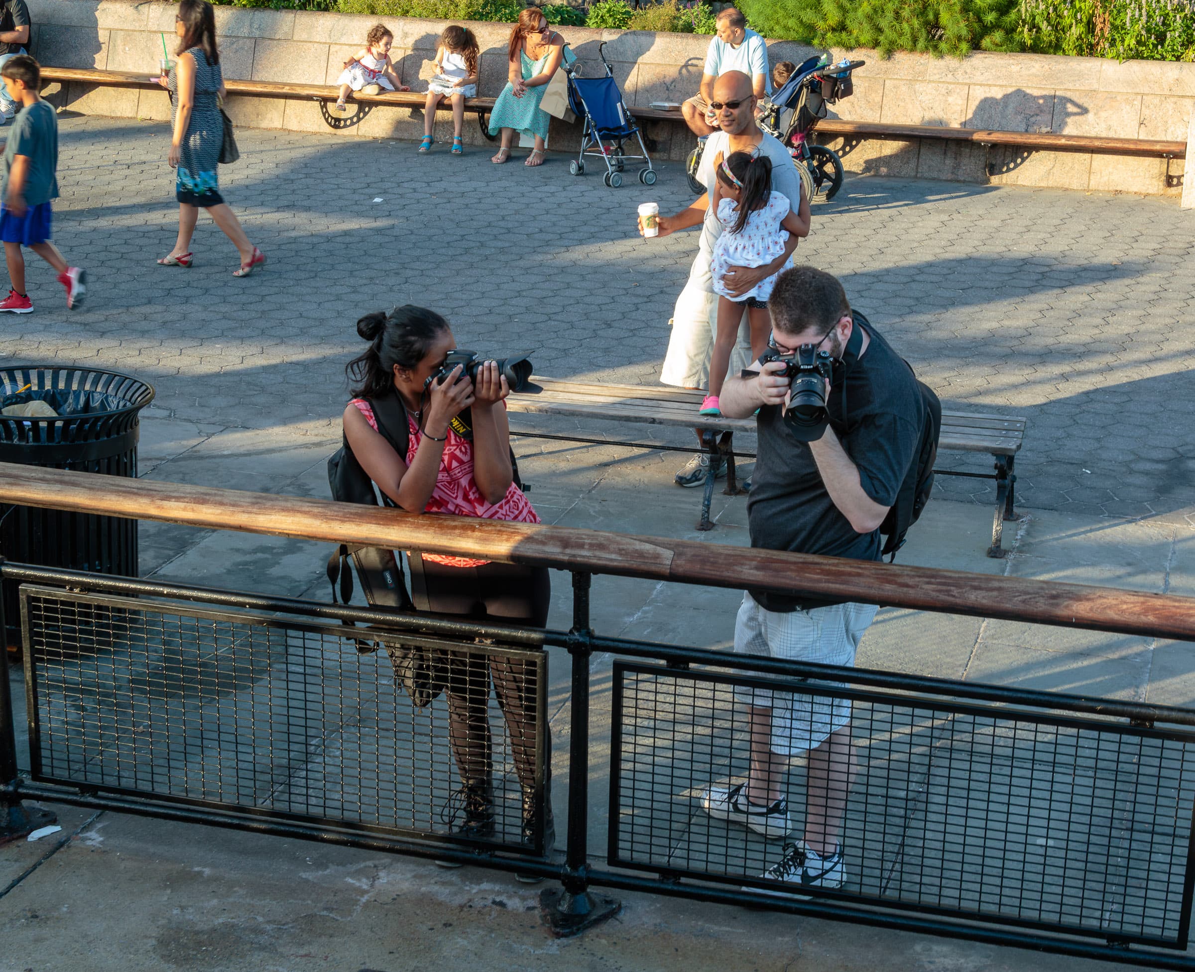 Two photographers concentrate on subjects outside the frame while people gather around them at The Battery in New York City in While the Photographers Were Looking by Francisco Colón.