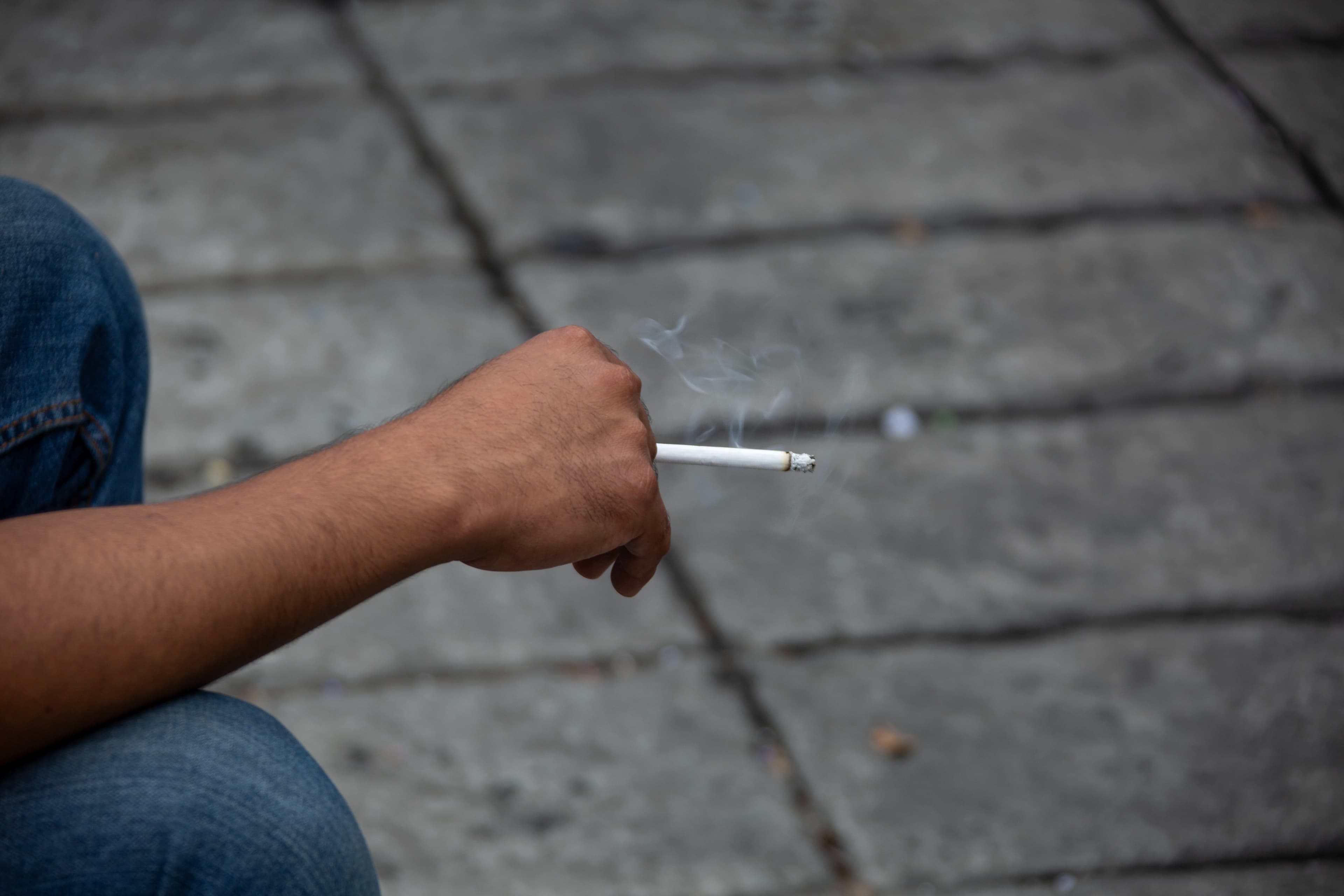 Close view of an unidentified seated person's arm and hand holding a cigarette as thin smoke rises against softly blurred pavement during the Mexico City Pride celebration.