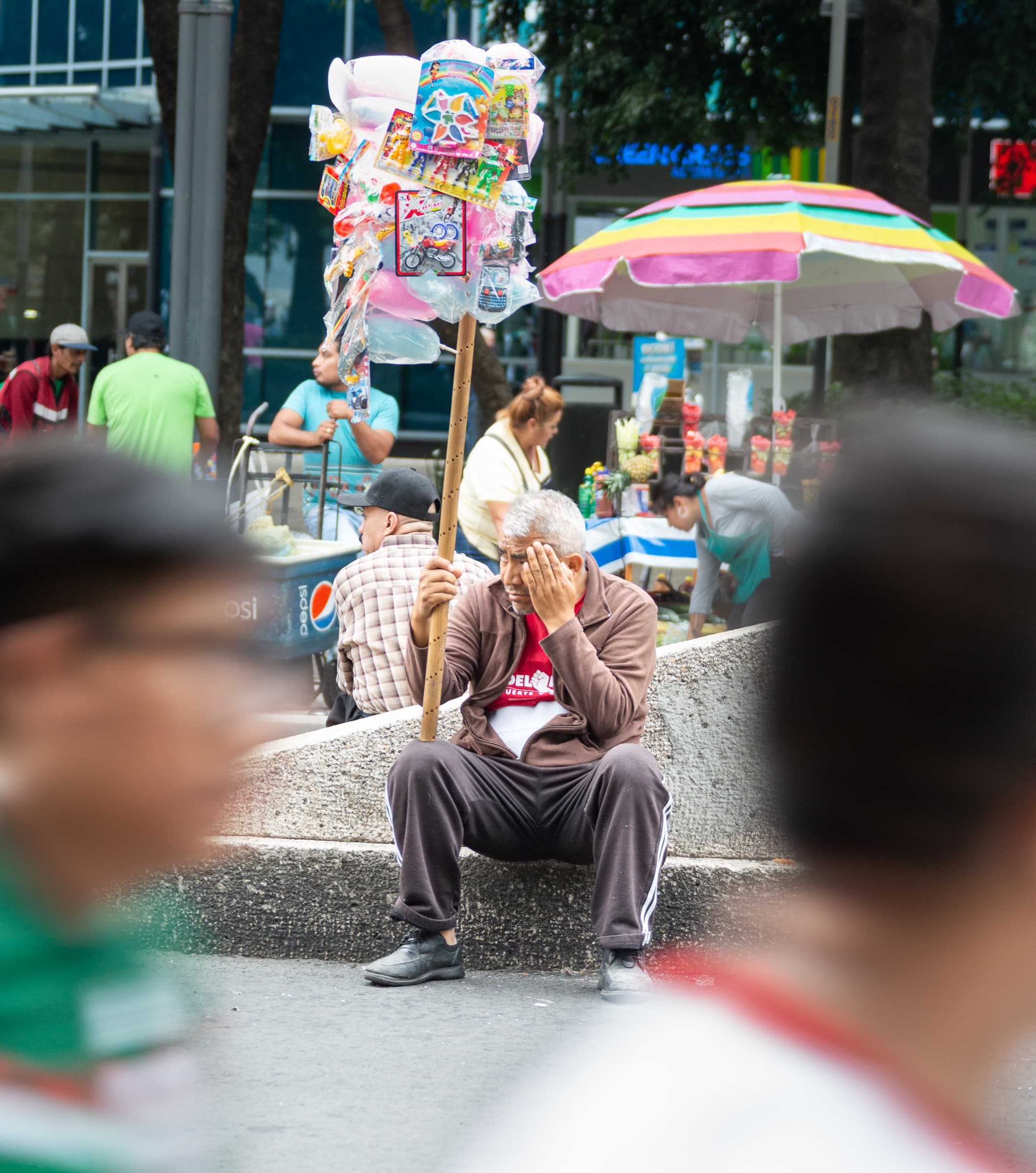 An older street vendor sits with one hand covering his face while holding a merchandise pole as blurred pedestrians pass during a public event in Mexico City.