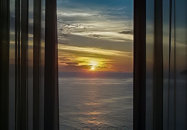 Atlantic sunrise photographed from inside a room at the Sheraton Hotel in Rio de Janeiro, framed by partially closed dark curtains and reflective window panels, with golden morning light extending across the ocean.
