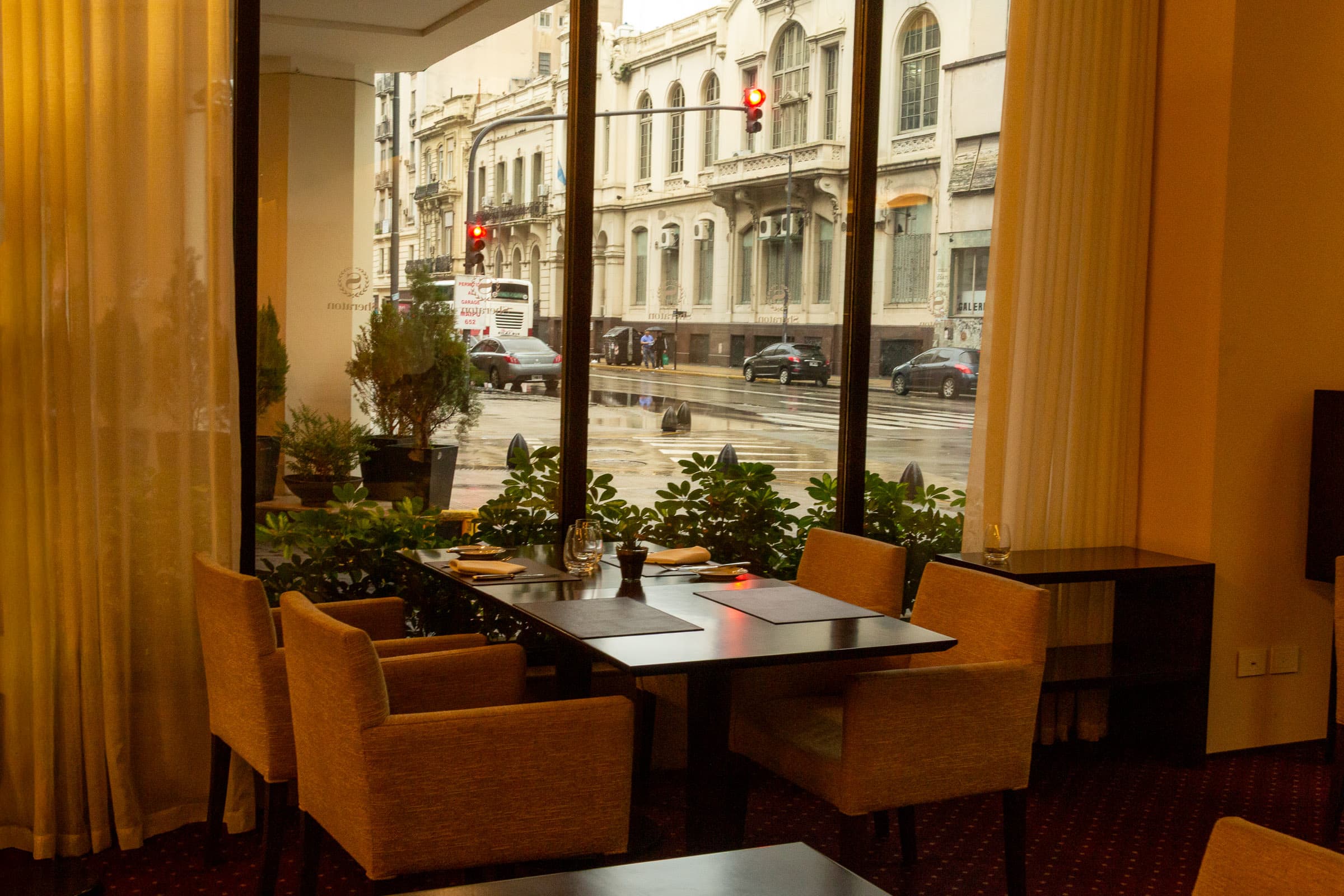 Warm hotel restaurant seating beside floor-to-ceiling windows overlooking a rain-soaked Buenos Aires street in On This Side of the Glass by Francisco Colón.