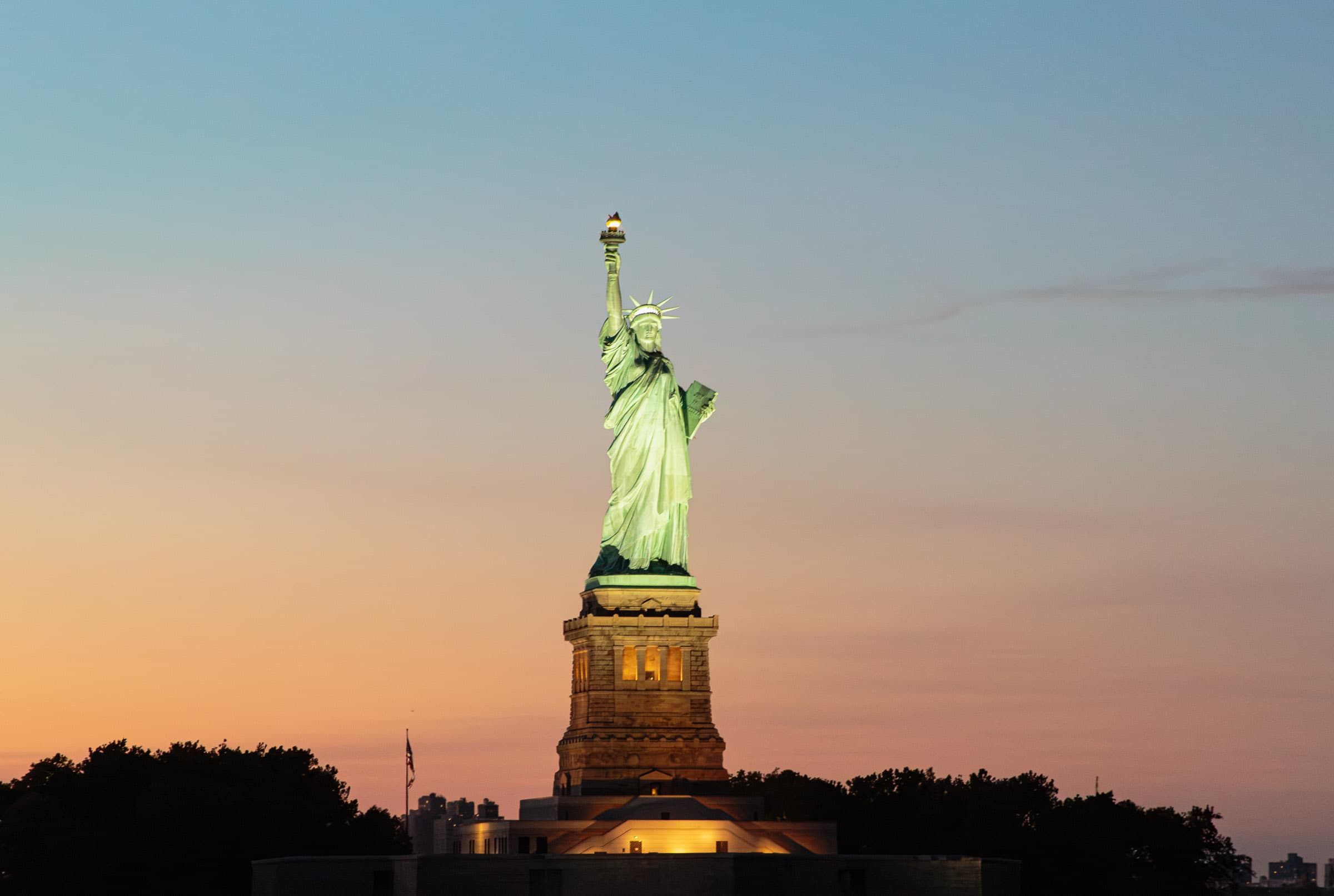The illuminated Statue of Liberty rises above Liberty Island against an evening sky transitioning from warm orange near the horizon to blue above in The Picture That Stayed by Francisco Colón.