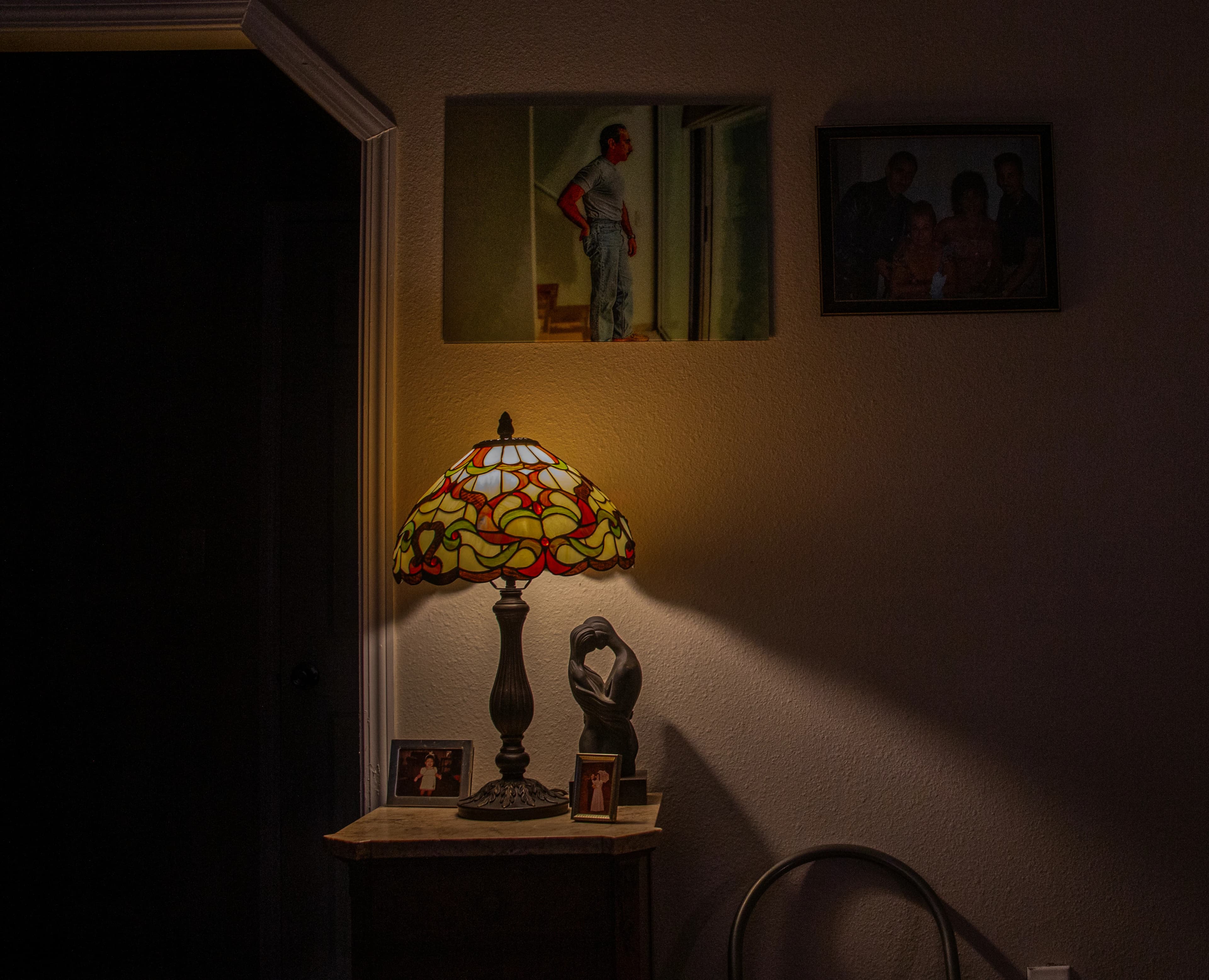 A stained-glass lamp illuminates framed family photographs, a self-portrait, and an embracing sculpture while a doorway and family portrait remain in shadow inside the artist’s home.