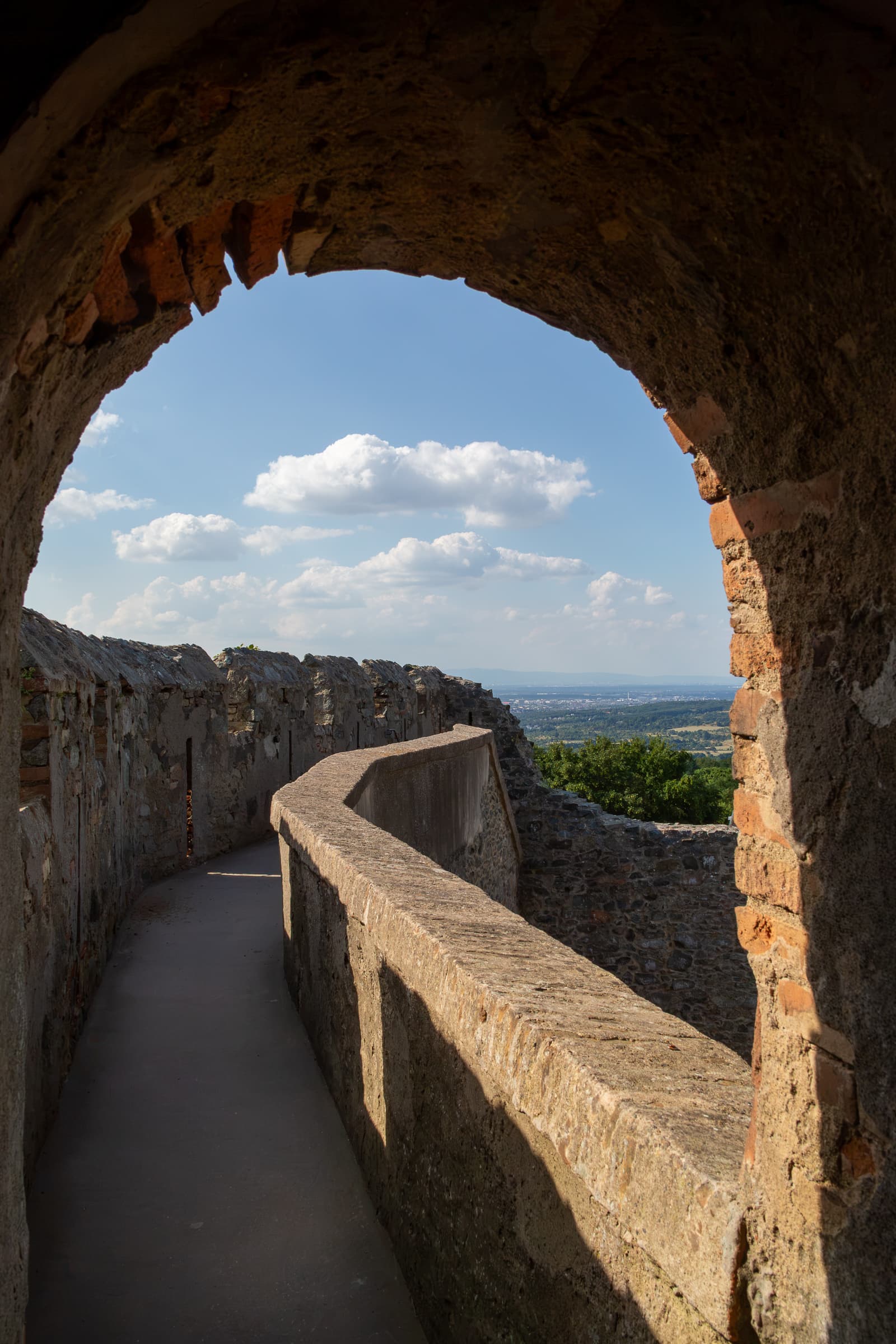 A curved stone walkway beneath a shadowed arch at Frankenstein Castle, opening toward a blue sky and distant landscape.
