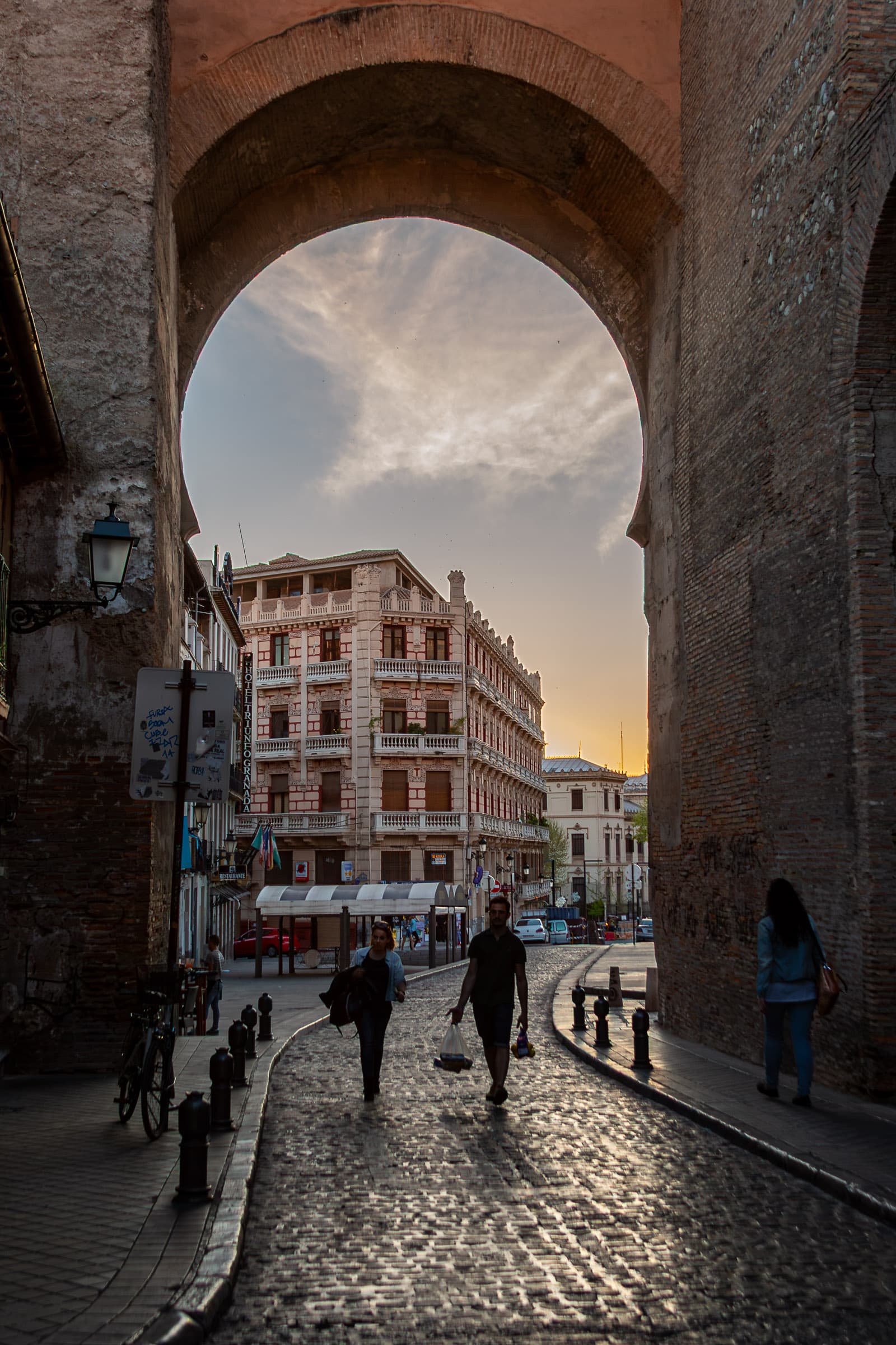 Pedestrians walk along a light-covered cobblestone street beneath a monumental historic arch in Granada, Spain, as evening light fades over the city.