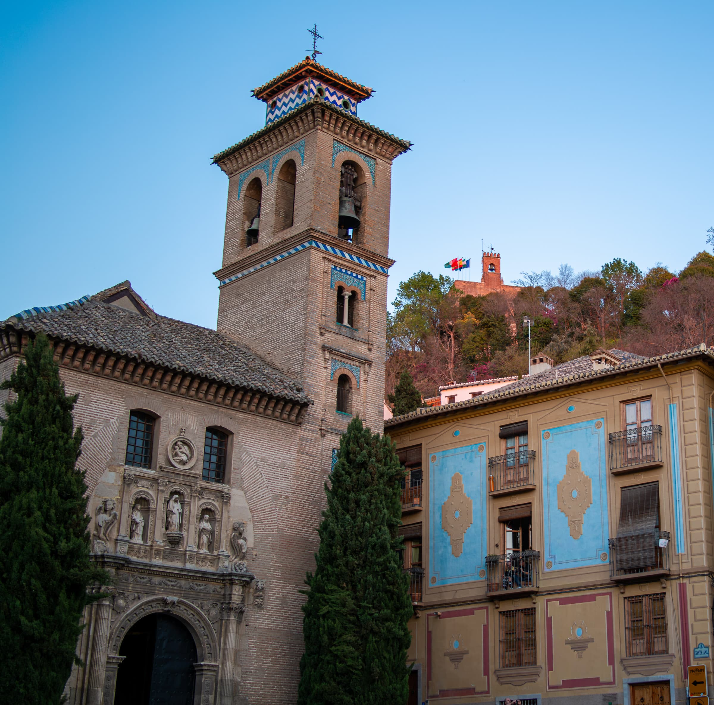 Iglesia de San Gil y Santa Ana beside an aqua-decorated building near Plaza Nueva in Granada, Spain, with two figures visible on a balcony and a distant tower on the hillside.