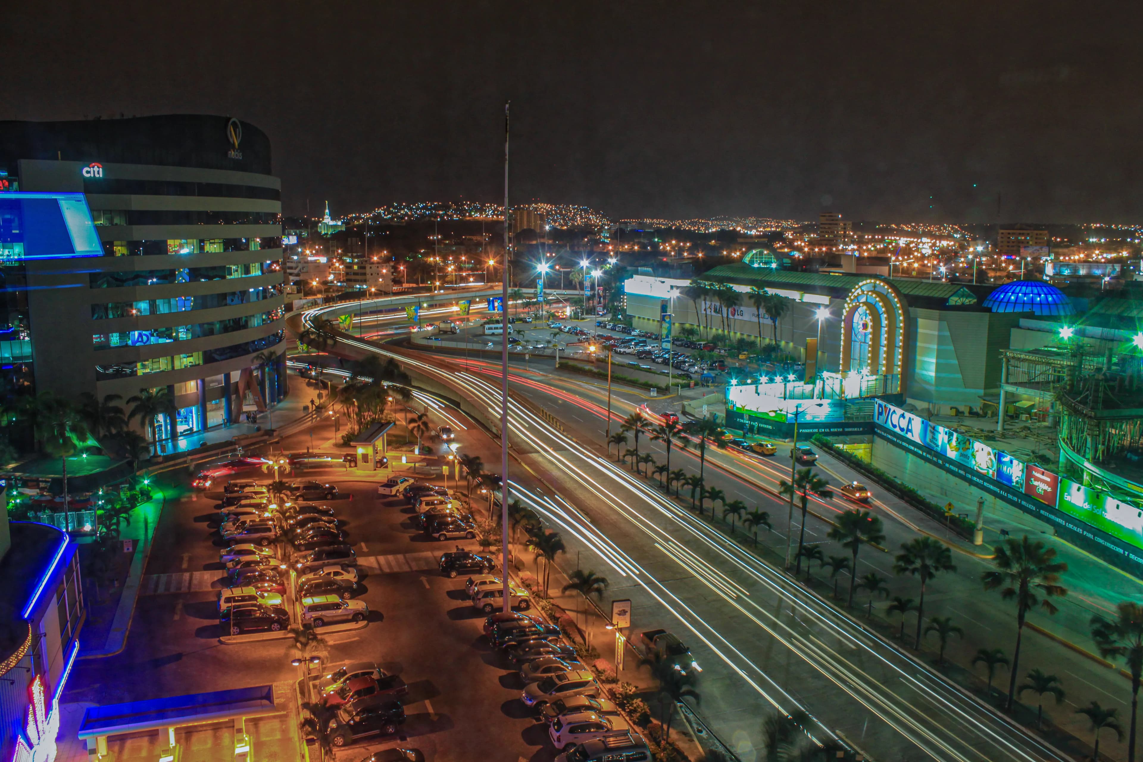 Night traffic in Guayaquil, Ecuador, creating white and red light trails along a curving roadway beneath illuminated city buildings, titled Guayaquil in Motion by Francisco Colón.