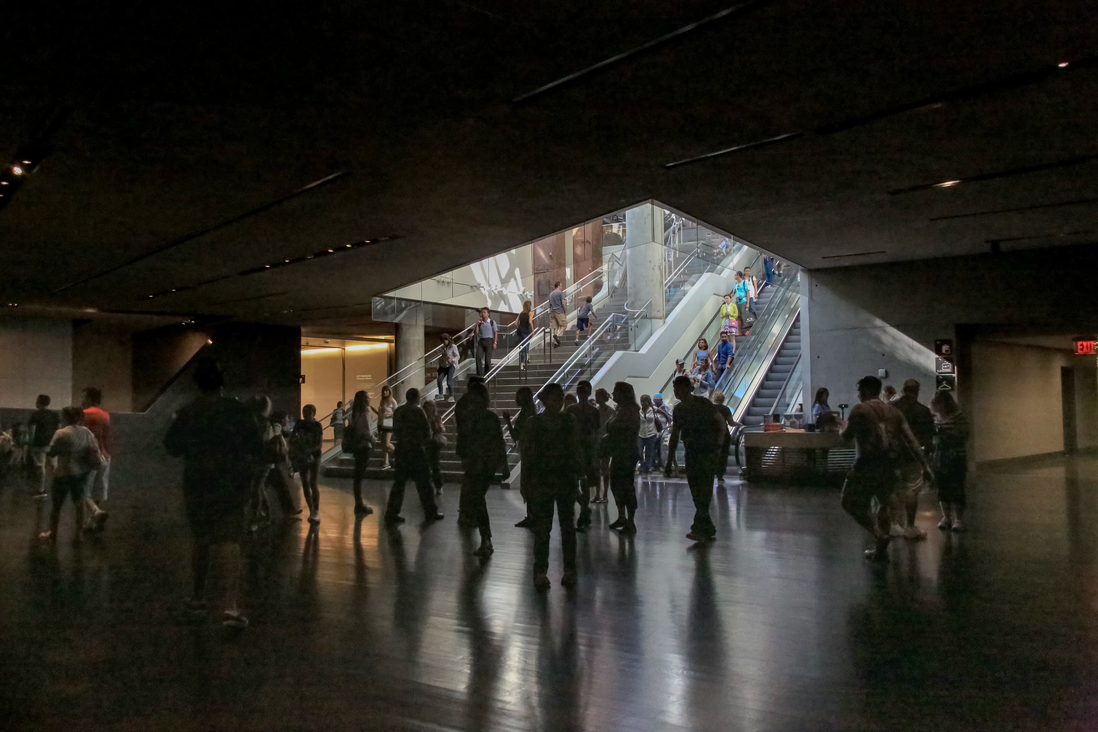 Visitors moving through the dark interior of the 9/11 Memorial and Museum in New York City, framed by monumental concrete architecture, stairs, escalators, and reflected light, titled Into the Memorial by Francisco Colón.