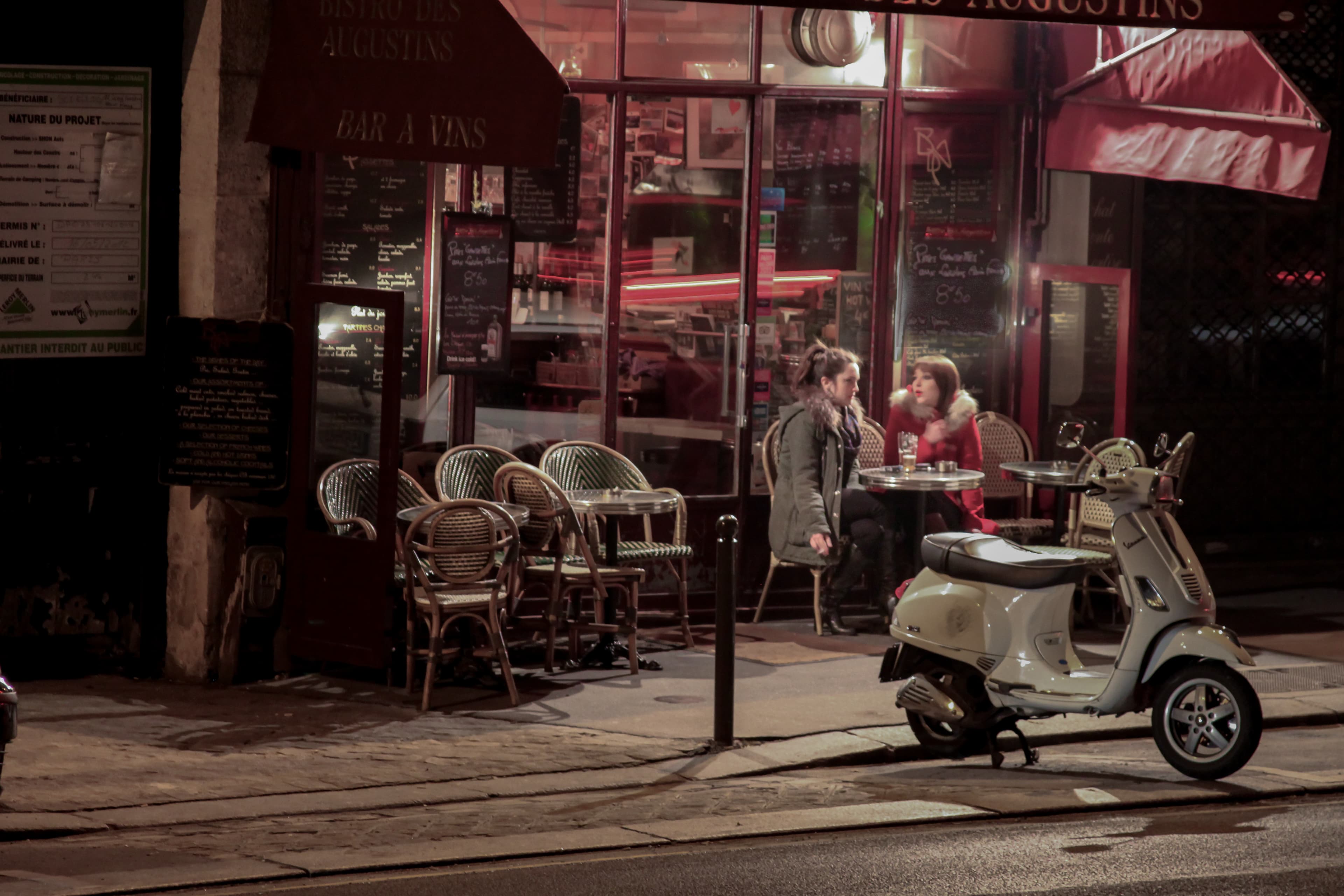 Two women converse outside a Paris café at night beside empty café chairs and a white scooter parked along the curb.