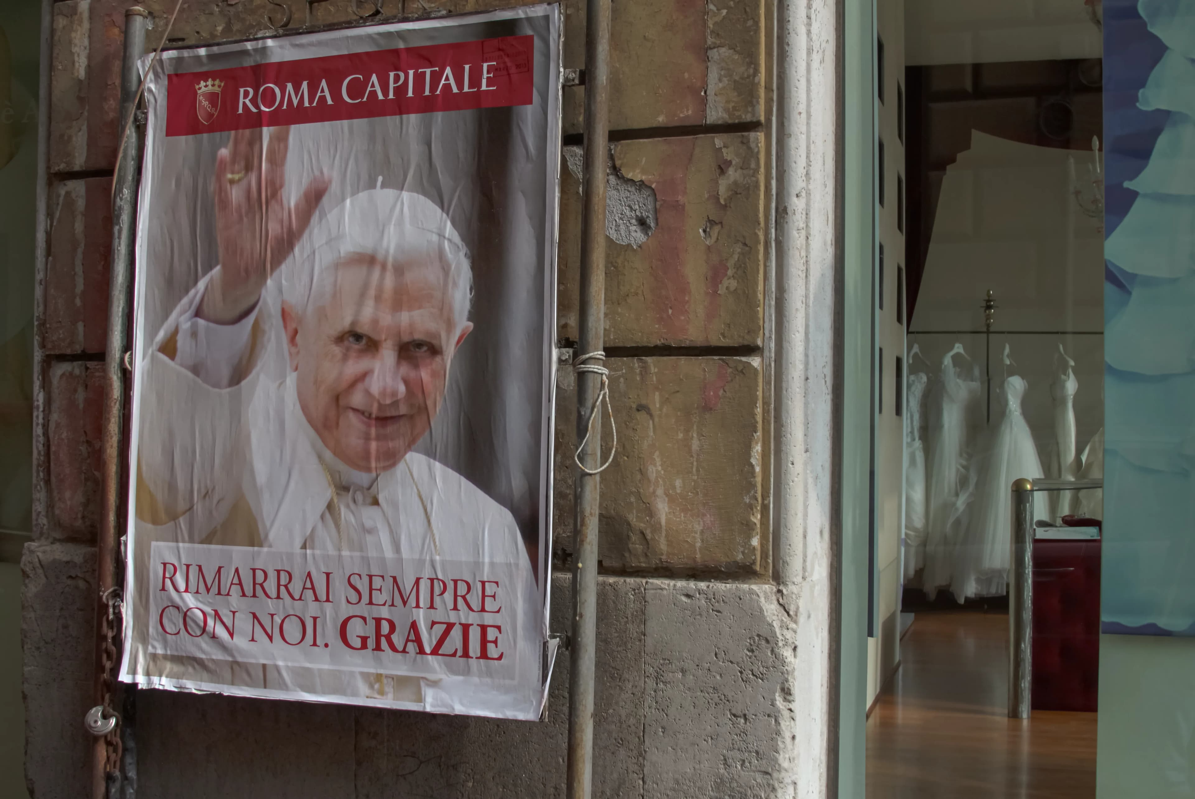Street poster of Pope Benedict XVI bearing the Italian words Rimarrai sempre con noi. Grazie on a weathered wall beside a storefront in Rome on February 28, 2013, titled Rome Says Goodbye by Francisco Colón.