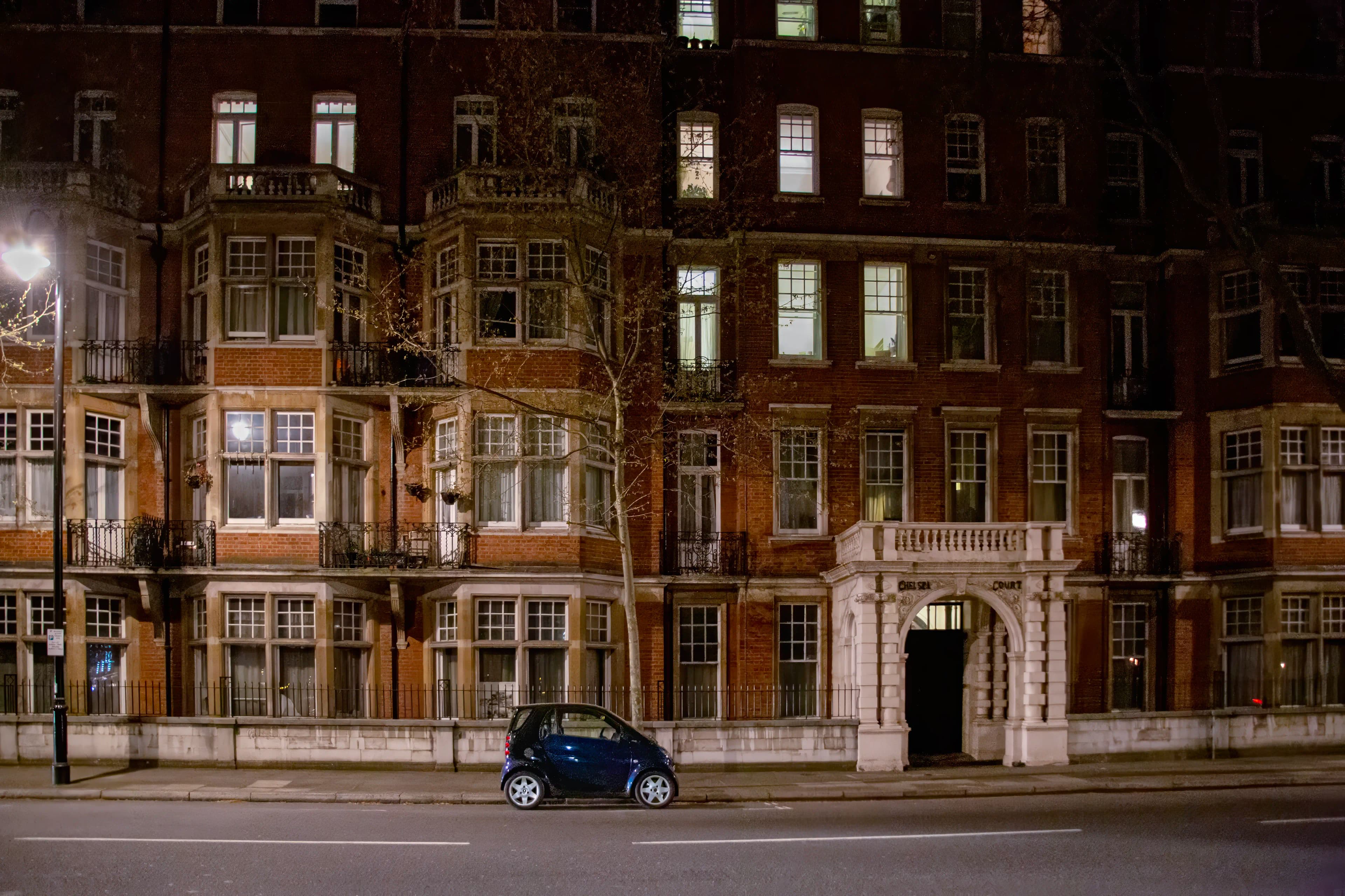 A solitary dark blue compact car parked beneath the towering red-brick façade and illuminated windows of Onslow Court in London at night.