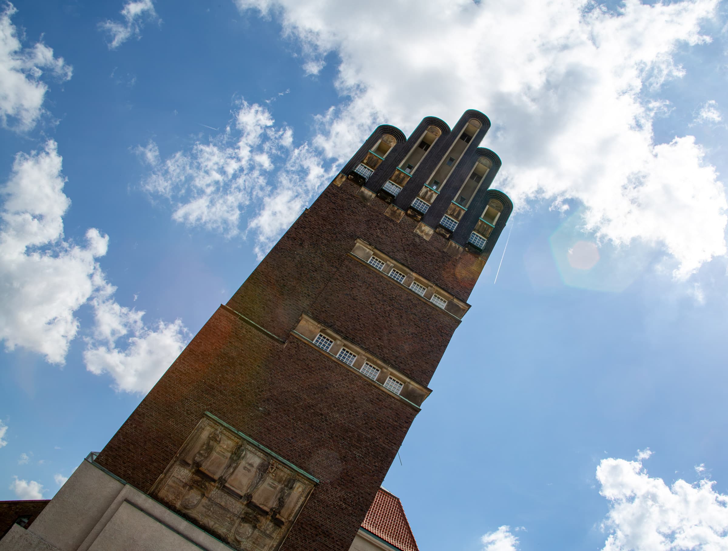 A dramatic low-angle view of Darmstadt's brick Wedding Tower rising diagonally into a blue sky filled with bright clouds and sunlight.