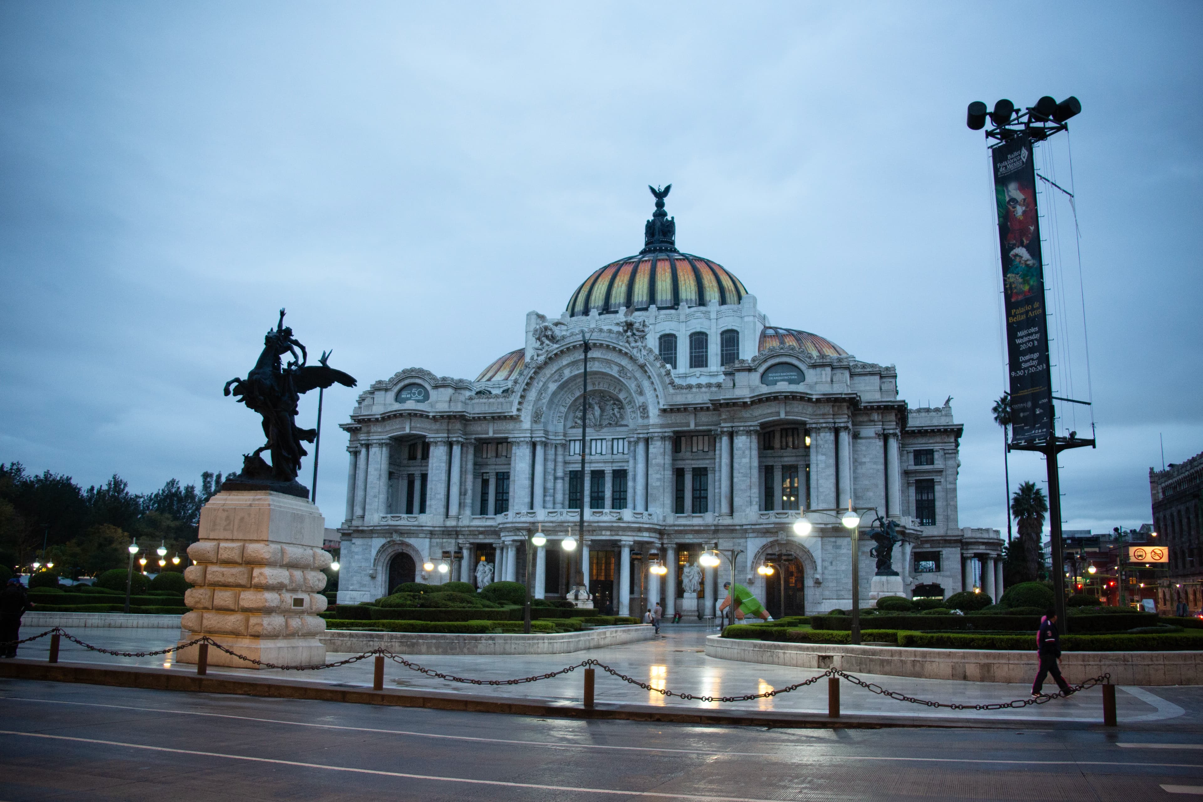 Palacio de Bellas Artes in Mexico City beneath a cool overcast morning sky after rain, with illuminated streetlights reflecting on wet pavement, a monument at left, a pedestrian at right, and contemporary city infrastructure surrounding the historic building.