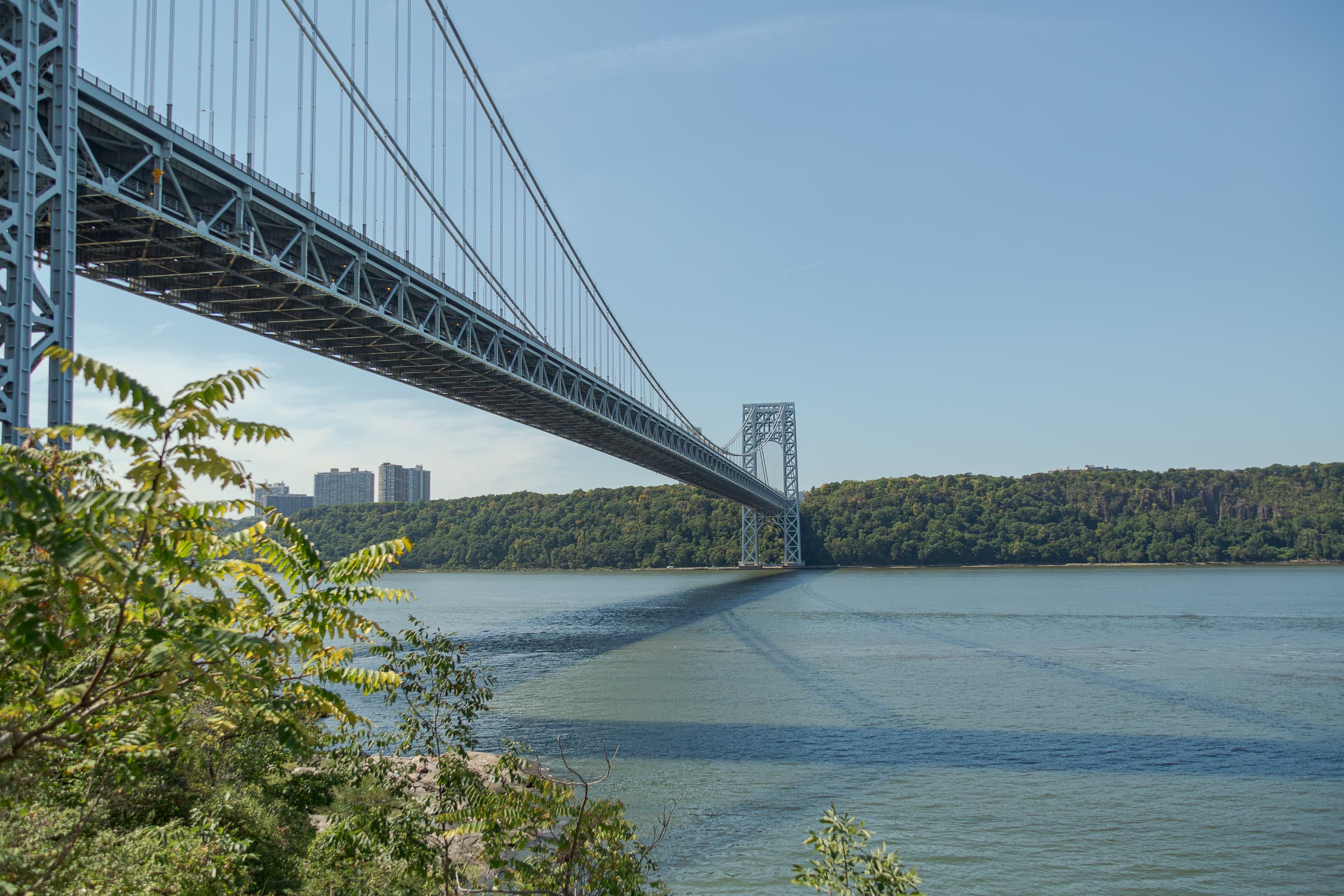 The George Washington Bridge extending diagonally above the Hudson River from a wooded path in Manhattan, with its shadow crossing the water toward Fort Lee and the New Jersey Palisades.