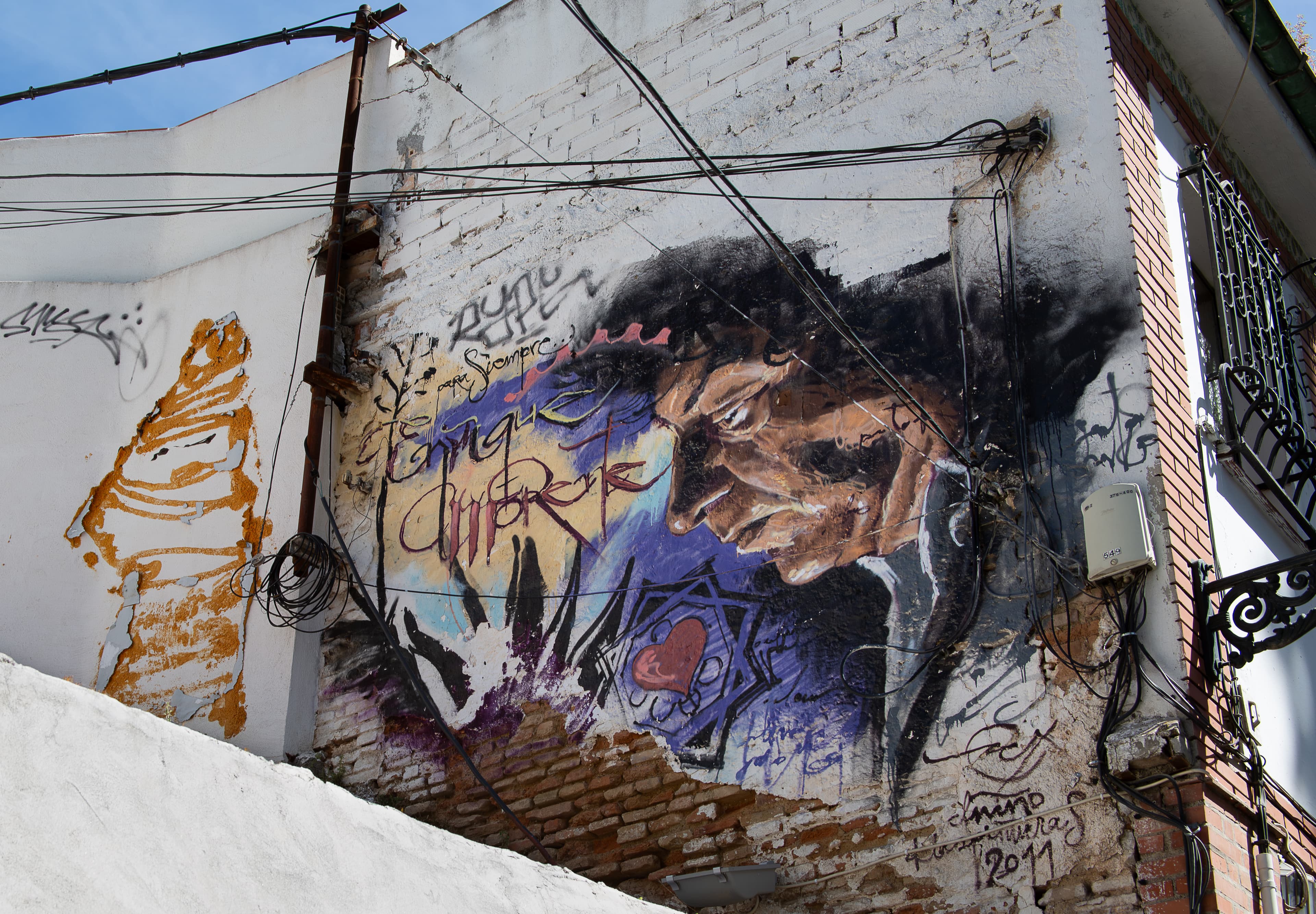 A painted portrait of flamenco singer Enrique Morente on a weathered Sacromonte wall, surrounded by graffiti, exposed brick, and crossing cables.